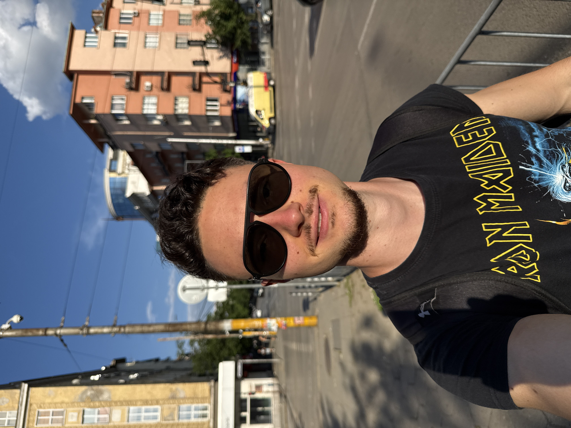 The media file is a selfie taken by a young man in Sofia, Bulgaria. The man is wearing a black t-shirt with a yellow "Iron Maiden" logo and graphic on it. He is also wearing sunglasses. The background shows a street scene with buildings, trees, and a yellow van. The time of day appears to be daytime, with bright sunlight, suggesting clear weather. The buildings in the background are typical of an urban environment, with a mix of older and newer architecture. The man's expression is neutral, and the overall mood of the image is casual. The photo is oriented vertically, and the man is looking towards the camera.