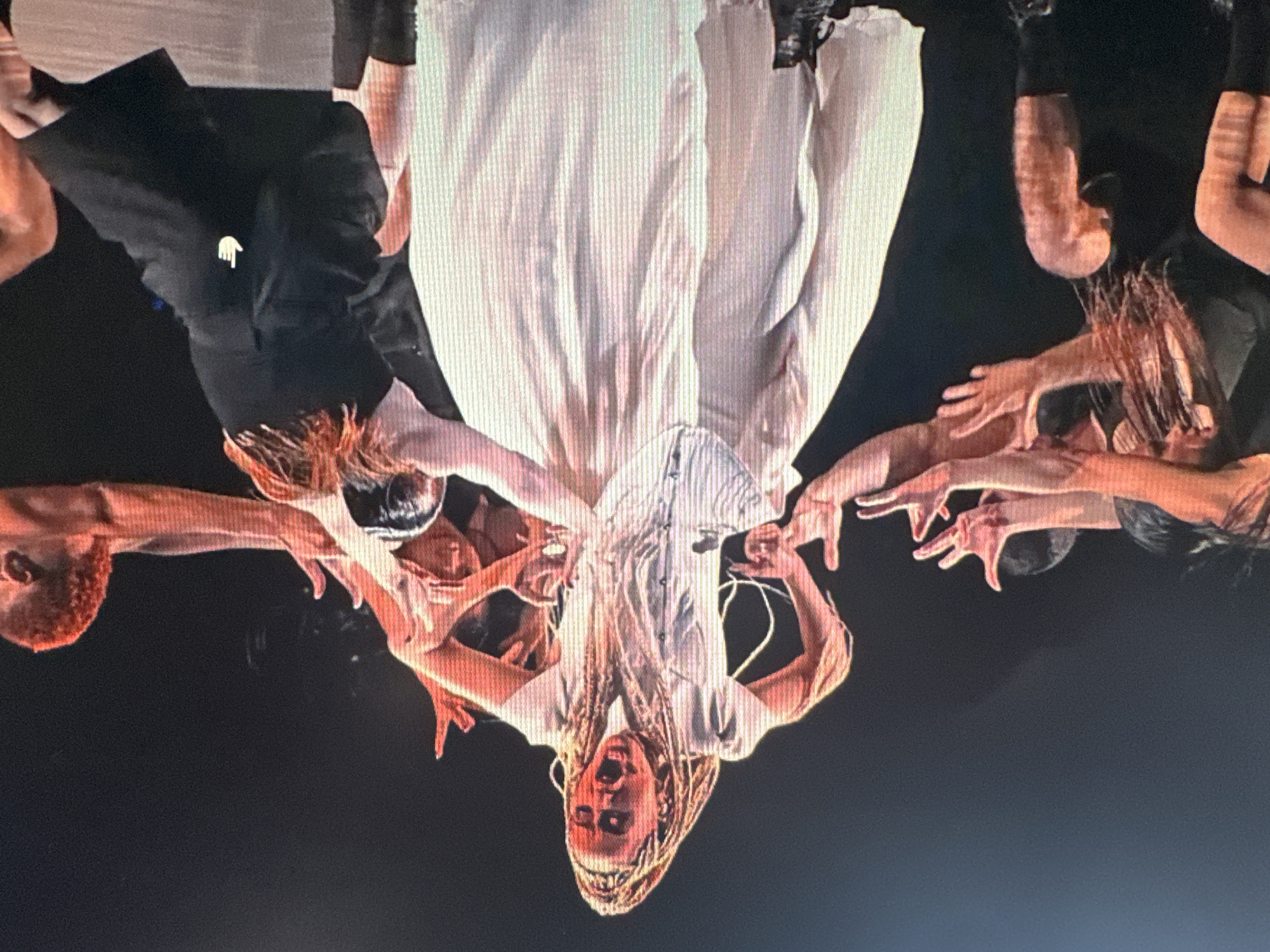 The image shows a performance on a stage with a dark background. In the center, a woman with blonde braided hair is wearing a white ruffled dress and a white corset. She is in a dramatic pose, with her mouth open in a shout and her arms outstretched. Surrounding her are several dancers in black attire, with their arms reaching towards her. The lighting on stage casts a reddish hue on some of the performers, particularly their skin and hair. This scene appears to be a live performance, likely a dance or theatrical production, but its specific location in Ellwangen, Germany, cannot be determined from the image alone.