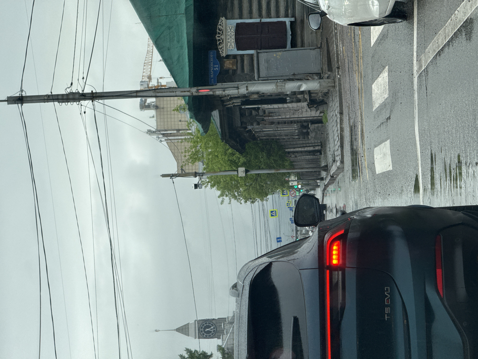 The image depicts a street scene in Krasnoyarsk, Russia on a cloudy day. The shot appears to be taken from inside a vehicle stopped in traffic. A black TS EVJ car is prominently visible. Buildings line the street, including one with a blue sign that reads "ул. Марковского, 51" (Ulitsa Markovskogo, 51), indicating the location on Markovskogo Street. A clock tower is visible in the distance. There are street signs and traffic signals along the street. Power lines and poles are visible above the street. The road is wet, suggesting recent rain. A crane is also visible in the background, indicating construction in the area. A white car is partially visible in the background to the right.