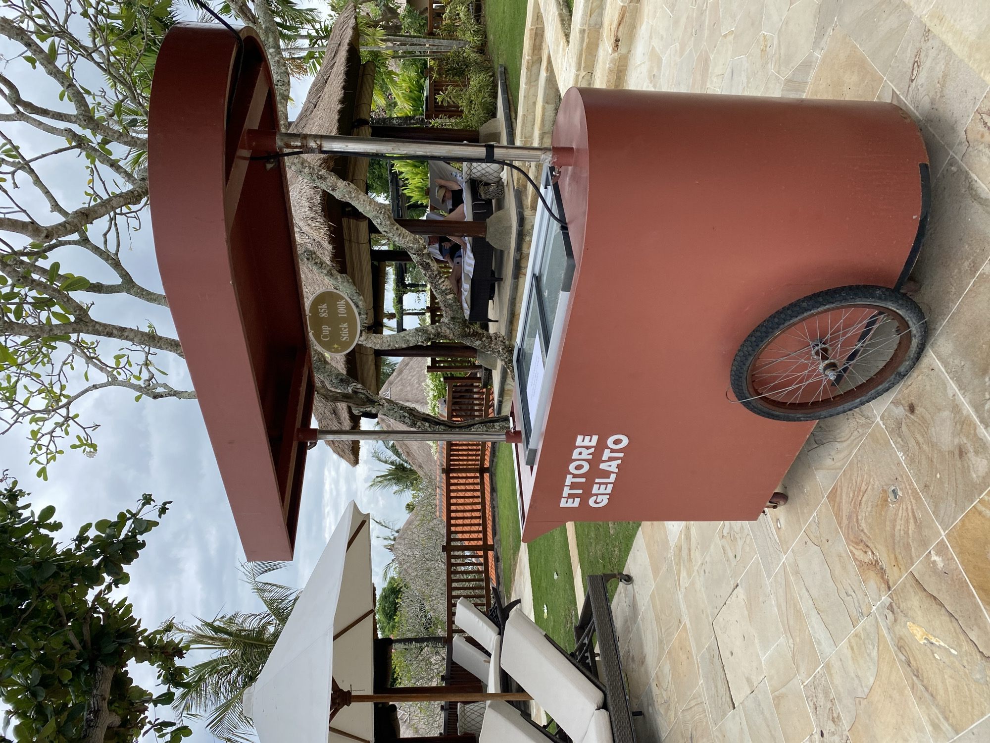 In Kuta Selatan, Indonesia, a red gelato cart with the name "ETTORE GELATO" is positioned outdoors on a stone-tiled patio. The cart has a display freezer and a single wheel on its right side. In the background, several white lounge chairs are visible on a grassy area, some beneath a large white umbrella. Further back, a thatched-roof pavilion houses two individuals relaxing on lounge chairs. The scene is surrounded by lush greenery and trees under an overcast sky.