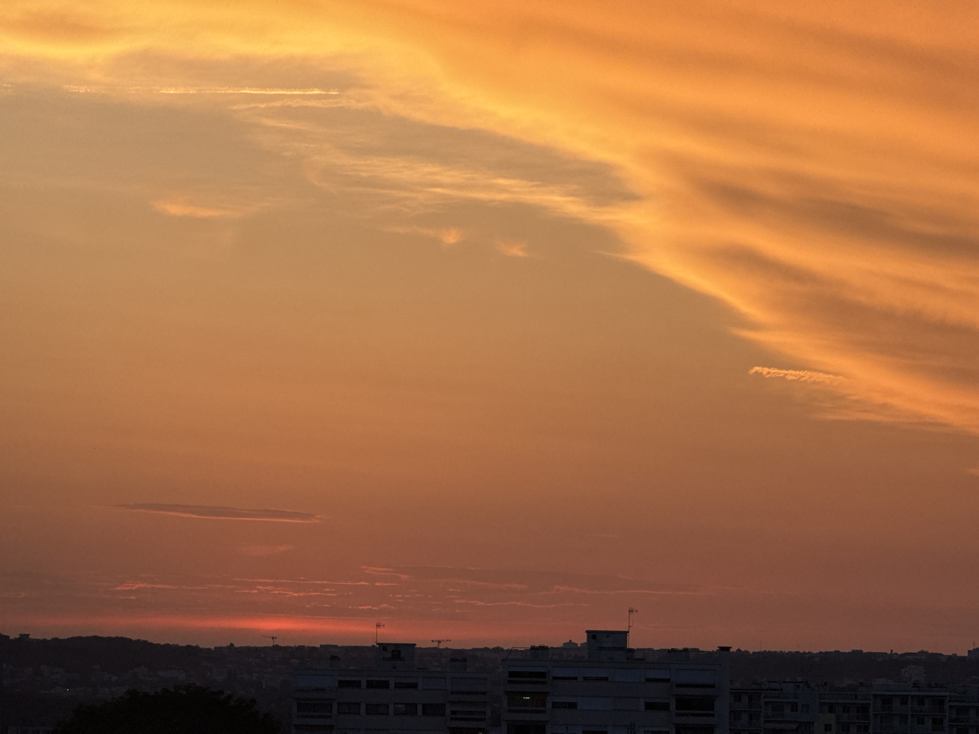 The image depicts a sunset over a city. The sky is filled with vibrant orange and yellow hues, with wispy clouds stretching across the upper portion of the frame. Below the sky, a silhouetted cityscape is visible, with buildings and trees forming a dark outline against the bright horizon. The overall scene evokes a sense of warmth and tranquility.
