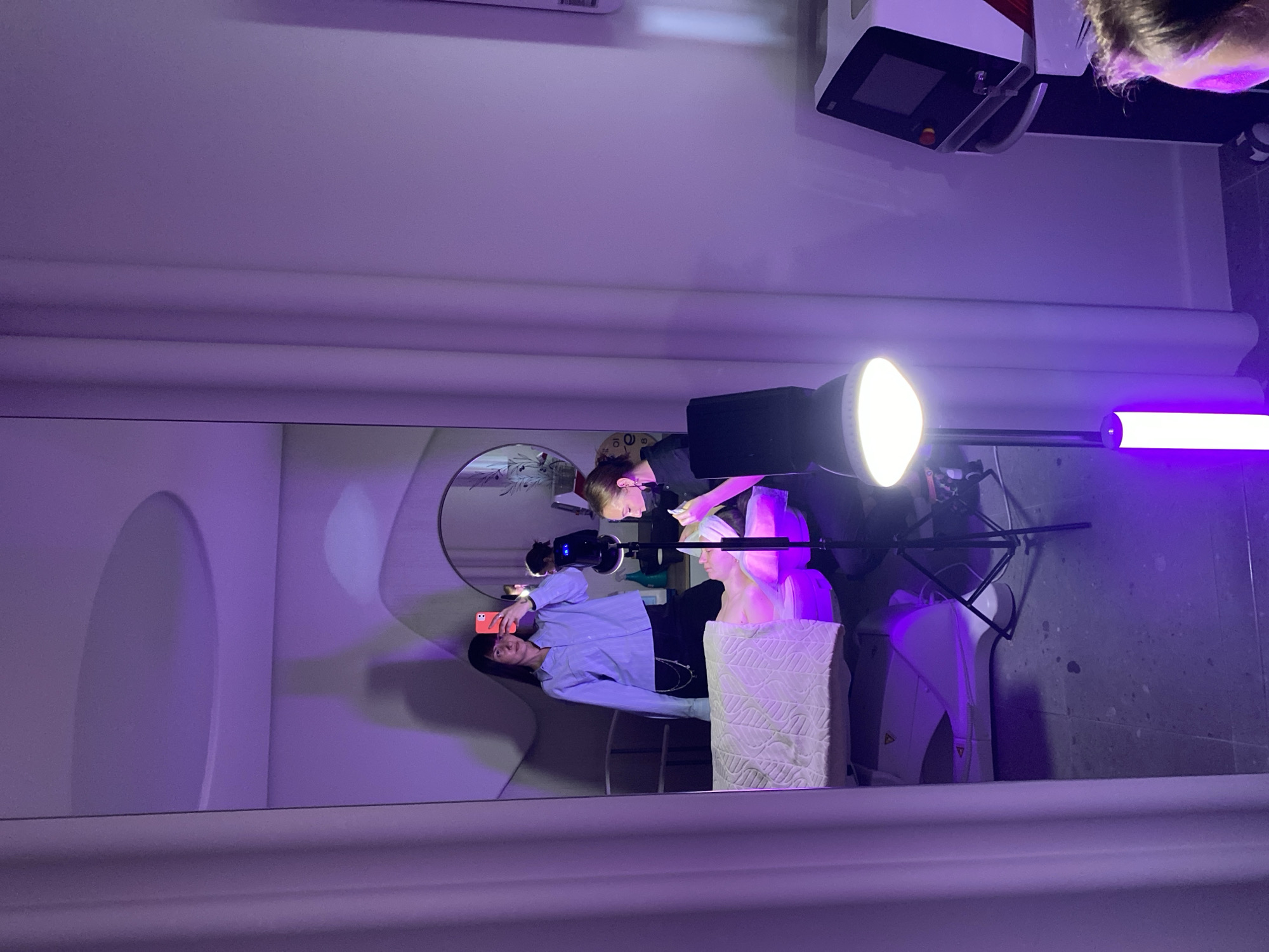 The image shows an indoor setting with purple lighting. A woman stands in front of a mirror, taking a picture with her smartphone. The mirror reflects her, a seated person receiving what appears to be a cosmetic treatment, and another individual, possibly a beautician, attending to the seated person. The backdrop features rounded wall elements, and professional lighting equipment is visible, including a ring light, a rectangular light, and an LED tube light. The scene suggests a beauty salon or treatment room. Location is Unknown, Unknown.