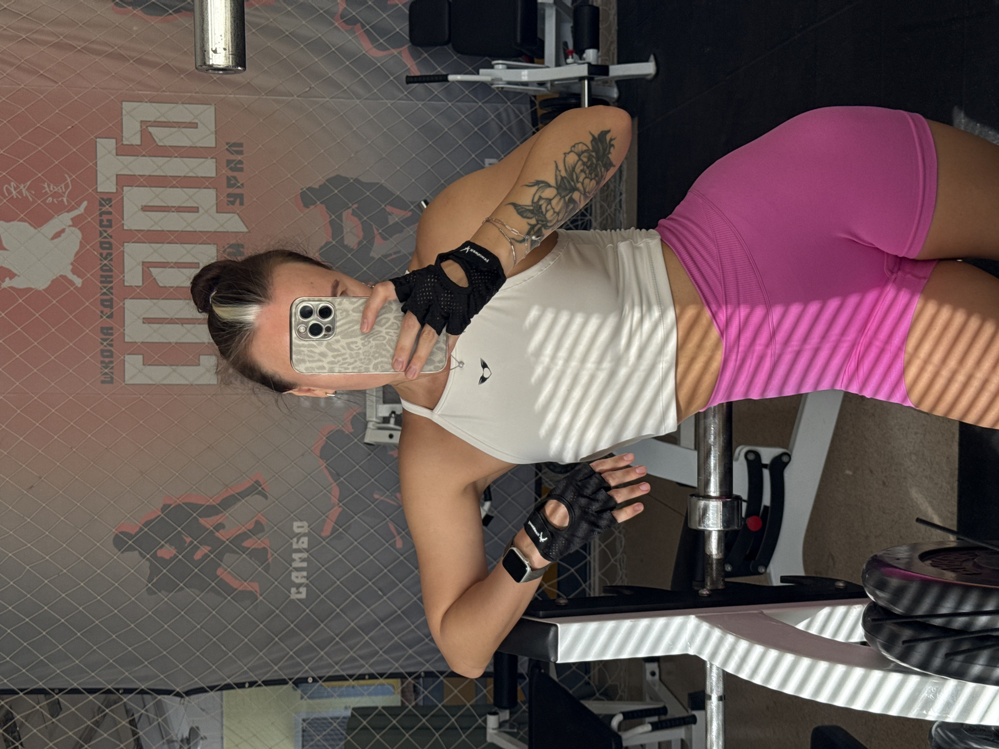 A woman is taking a selfie in a gym, possibly located in Chaikovsky, Russia, given the Cyrillic text and the "CAMEO" branding on the wall graphic behind her. The woman has her hair in a bun with a streak of blonde, wears pink athletic shorts, a white tank top, and black workout gloves. She is holding a smartphone with a patterned case, obscuring her face. Her left arm is visible, featuring a floral tattoo.
The background includes a chain-link fence and gym equipment like a leg press machine with weight plates. The flooring appears to be gray with a crosshatch pattern. The lighting conditions are somewhat bright, creating shadows.