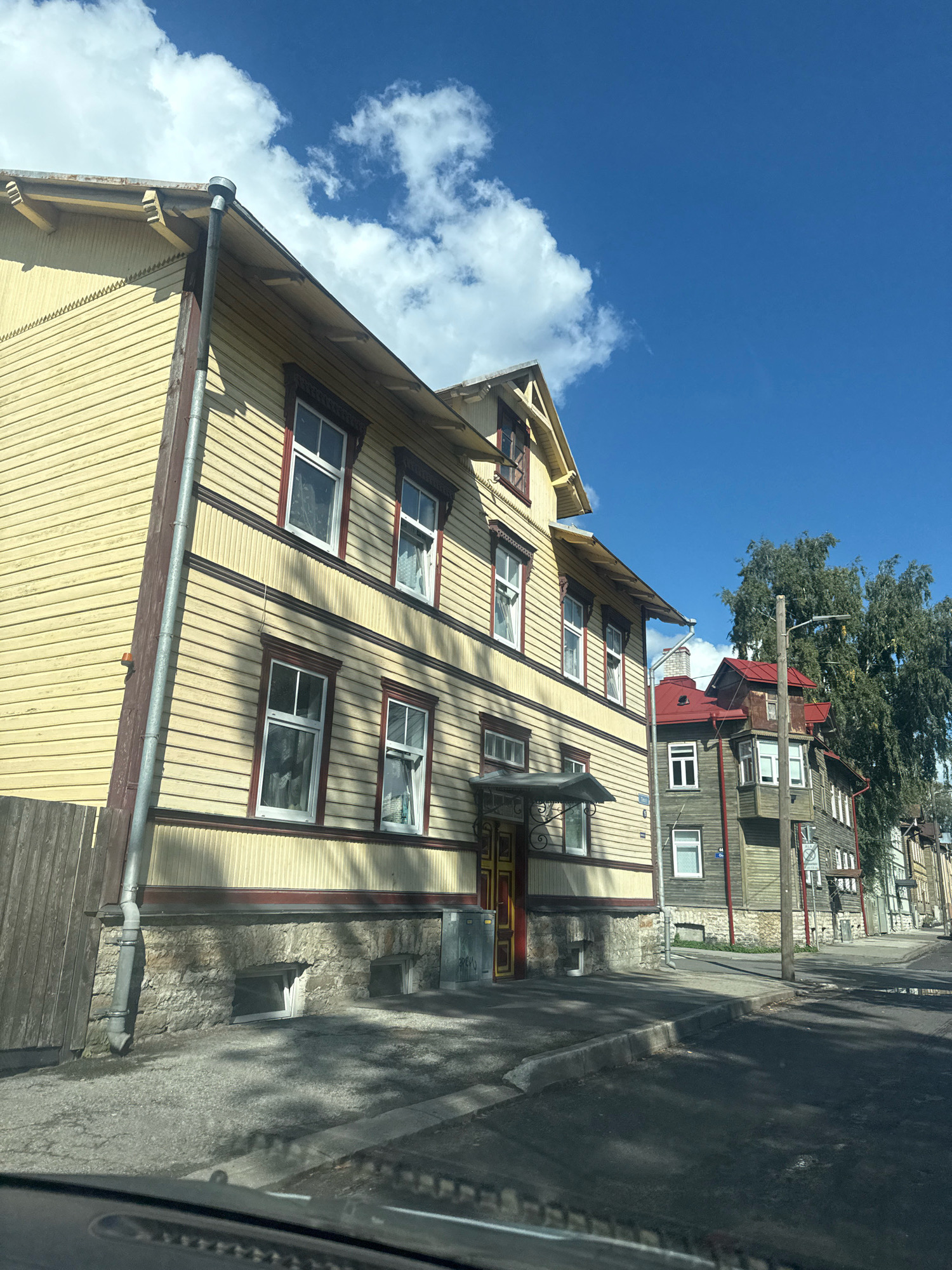 The image shows a street scene in Tallinn, Estonia. The foreground is dominated by the interior of a vehicle, with the dashboard visible at the bottom. The scene opens up to a street lined with traditional wooden houses, typical of areas like Kalamaja or Kadriorg in Tallinn. The houses are of varying heights, with the closest one being a prominent, pale yellow structure with intricate dark red trim around the windows and roofline. This house features a stone foundation with small basement windows. Next to it, a neighboring house has a dark green facade and a bright red roof. Further down the street, more houses with wooden siding and varied roof colors are visible, interspersed with trees. The sky is a clear blue with scattered white clouds. The street is paved, and a sidewalk is visible in front of the houses. There are no people visible in the image. The lighting suggests it is daytime, with sunlight casting shadows on the street and houses.