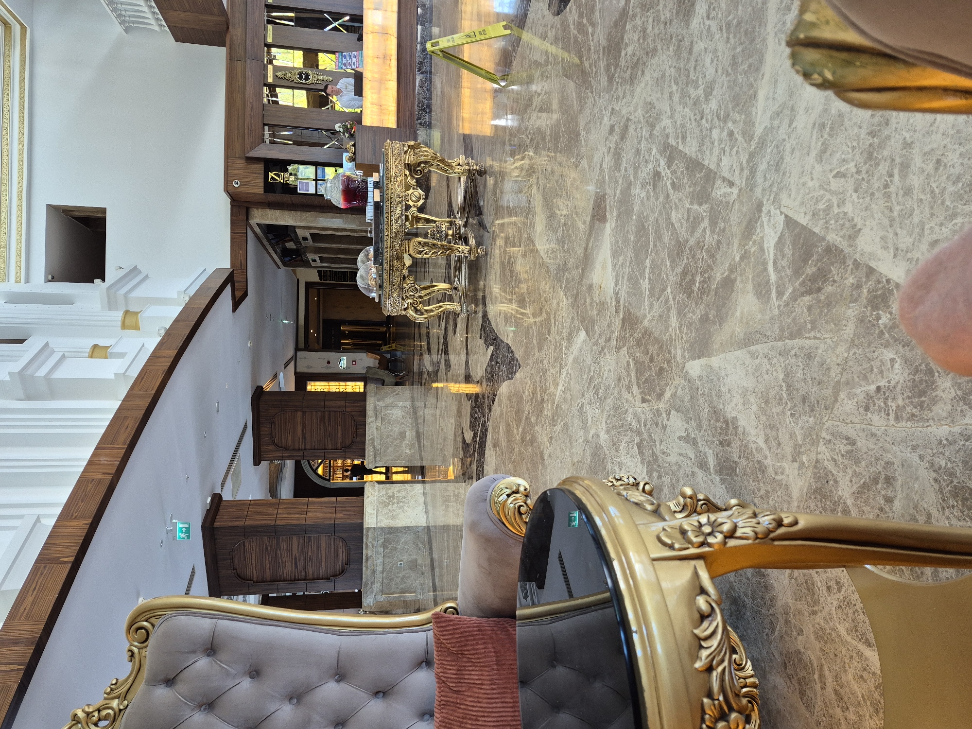 The image depicts an interior space, likely a hotel lobby in Alanya, Turkey, characterized by opulent decor. The flooring is marble with intricate patterns. In the foreground, there's a portion of a tufted velvet chair and a glass-topped table with a gilded frame. Further back, a decorative console table laden with beverages and ornate displays is visible near an open staircase with wooden trim and white risers. Beyond the table is a section of a hallway. A yellow safety sign is partially visible. The image suggests a luxurious and well-maintained environment.