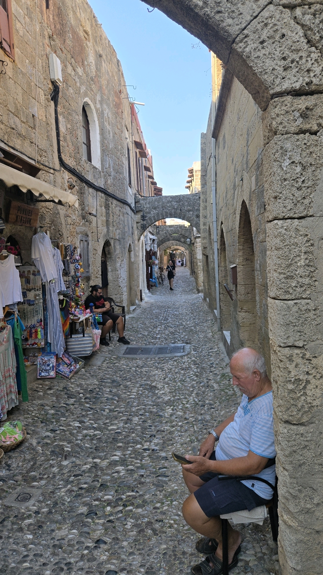 The image depicts a narrow, cobblestone street in the Municipal Unit of Rhodes, Greece. The street is lined with ancient stone buildings, some of which have arches spanning across the street. On the left side of the street, a shop displays clothing and souvenirs. A man is seated on a chair in front of the shop, looking at his phone. Further down the street, another person, appearing to be a woman, is walking away from the camera. In the foreground, an older man is seated on a stool, also looking at his phone. The sky above is clear and blue. The scene suggests a warm, sunny day in a historic urban environment.