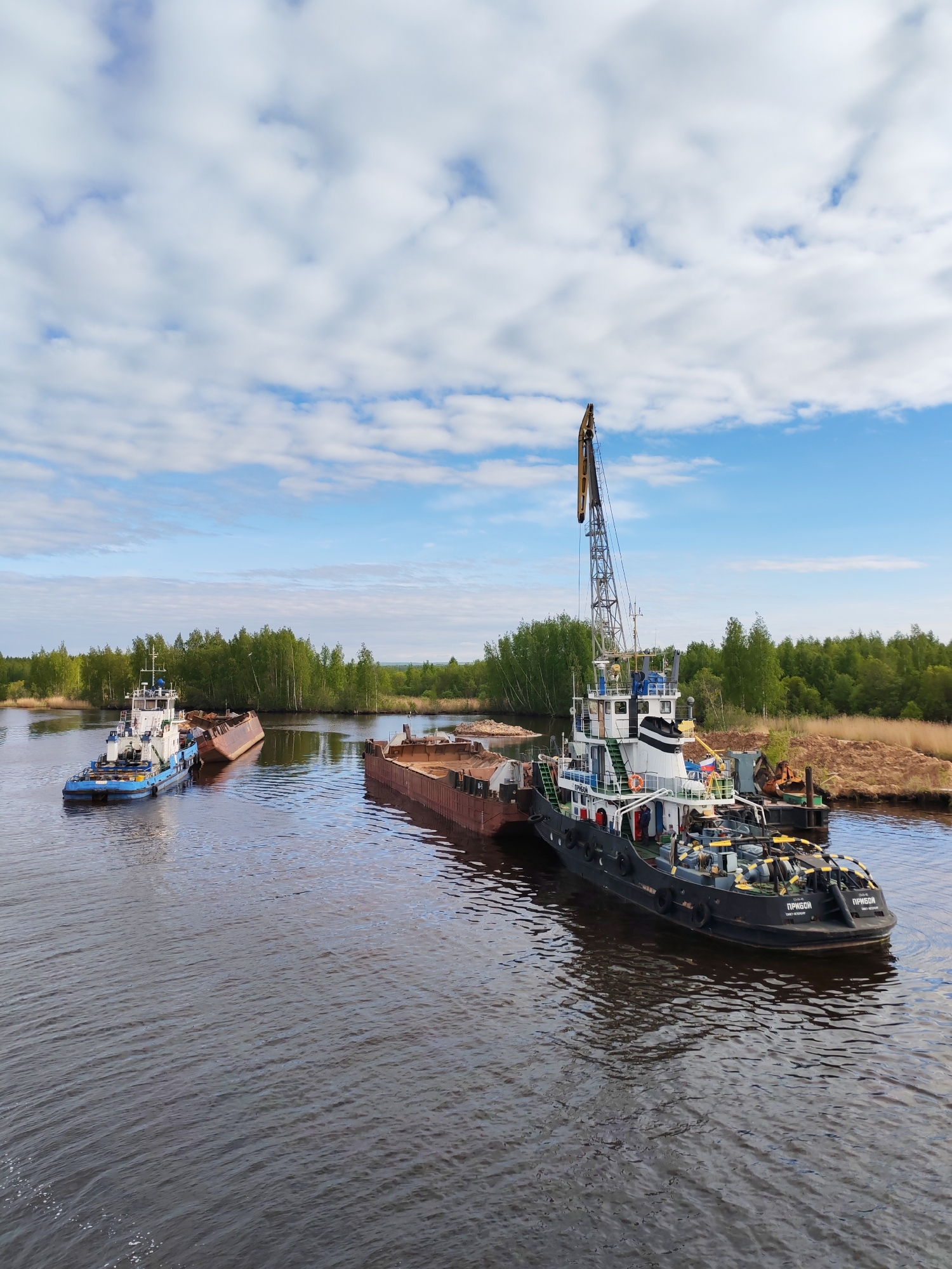 The scene depicts a waterway in Ozernoe Ust'e, Russia, with two tugboats and barges. The larger tugboat, primarily black with white accents, is equipped with a crane and is docked alongside a barge loaded with what appears to be sand or gravel. The tugboat has the inscription "ПРИБОЙ" on its hull. A smaller, blue and white tugboat is visible in the distance, towing another barge which seems partially submerged. The waterway is surrounded by green trees and vegetation on both banks, suggesting a rural or natural environment near an industrial operation. The sky is blue with scattered white clouds, indicating a fair weather day.
