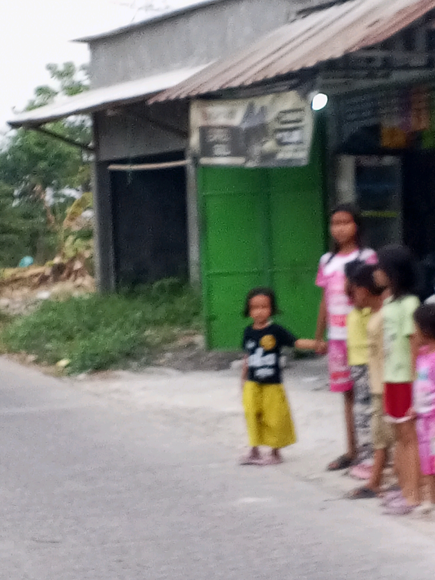 In Kadikaran, Indonesia, a blurry image captures a group of five young girls standing on an asphalt surface near a building with a corrugated metal roof. The building appears to be a shop or small establishment, with a green wall and a partially visible sign. The girls are positioned in a line, facing towards the right side of the frame. The girl closest to the left is wearing a black t-shirt with a yellow graphic and yellow pants. To her right, a girl in a pink and white striped t-shirt and pink and white striped shorts stands. Next to her, a girl in a yellow shirt and patterned pants. Further to the right, a girl wears a pale green shirt and red pants, and the girl on the far right is in a pink shirt and pink shorts. The ground in front of the girls is a paved area that transitions into a road. Lush green vegetation is visible behind the building. The overall lighting suggests an overcast day.