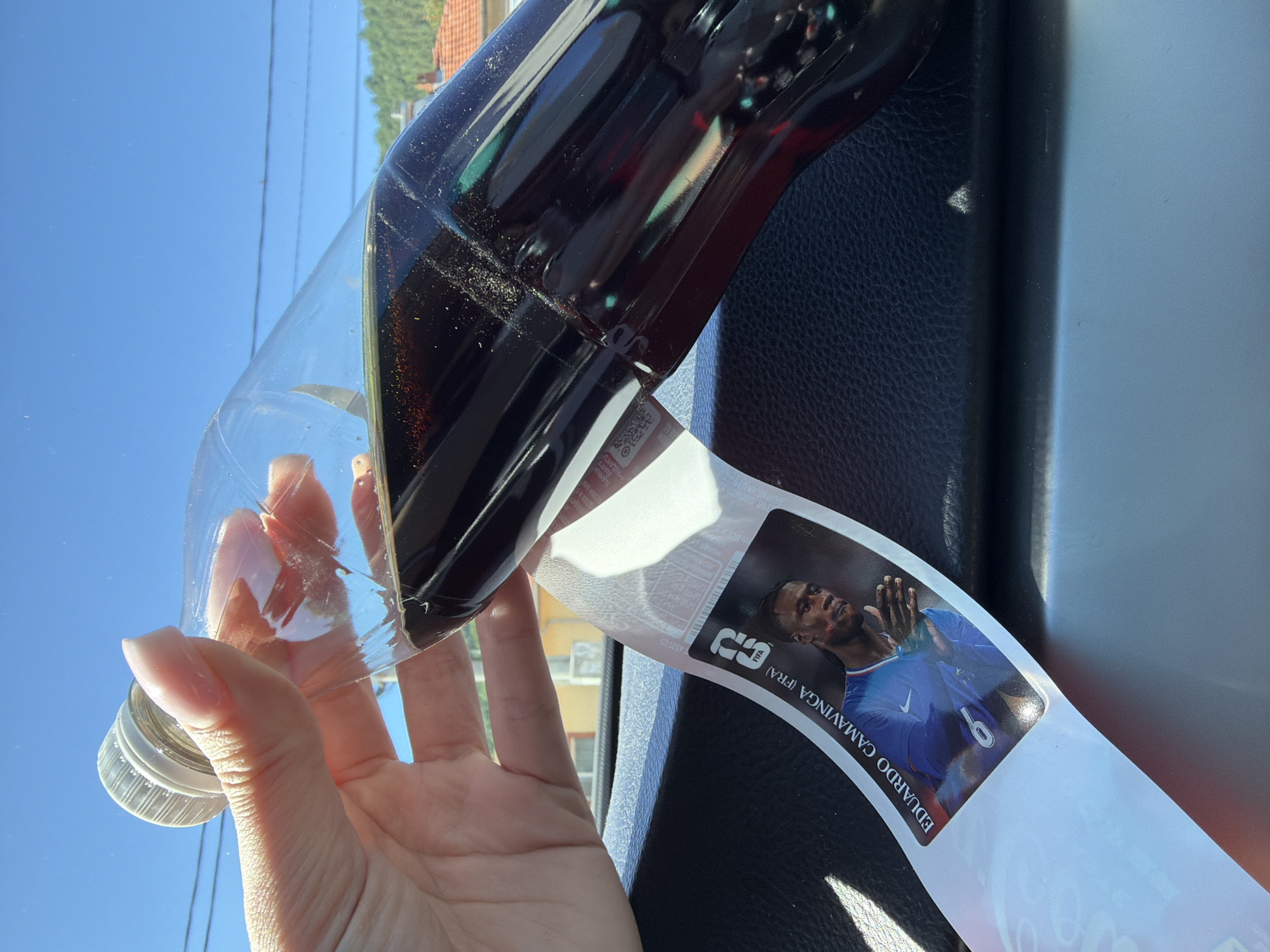 A hand holds a partially filled plastic bottle of dark soda, tilted forward, with the bottle's label featuring a picture of a soccer player, Eduardo Camavinga (FRA). The bottle is positioned over a black textured surface, likely a car's dashboard. In the background, a bright blue sky is visible with subtle power lines and distant greenery with red-tiled roofs, suggesting an outdoor, possibly urban or suburban setting. The image does not contain information indicating the specific location within the city of Beli Osam, Bulgaria.