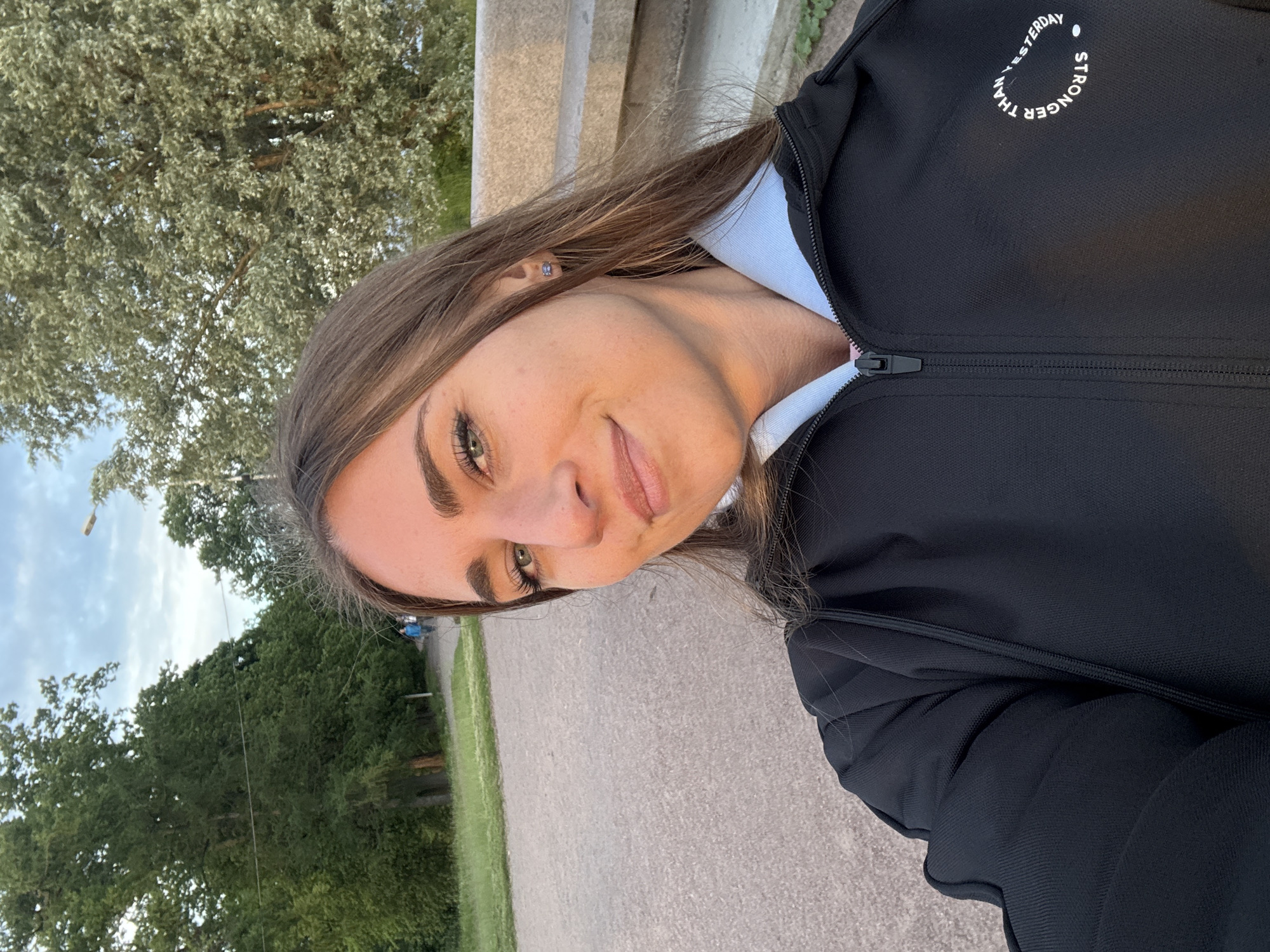 A young woman is taking a selfie outdoors. She is wearing a black zip-up jacket over a white shirt, and a small silver earring is visible in her left ear. The jacket has white text printed on the upper chest, which reads: "STRONGER THAN YESTERDAY". The woman has long brown hair and is looking at the camera with a slight smile. Her makeup is subtle, highlighting her green eyes and eyebrows.
The background suggests an outdoor setting, possibly a park or a pathway. There are trees with green foliage and a cloudy sky visible. The lighting indicates it might be late afternoon or early evening, with the sun casting a warm glow on the woman's face. A paved path runs through the scene. The image's orientation is rotated, with the woman's head appearing on the right side of the frame.