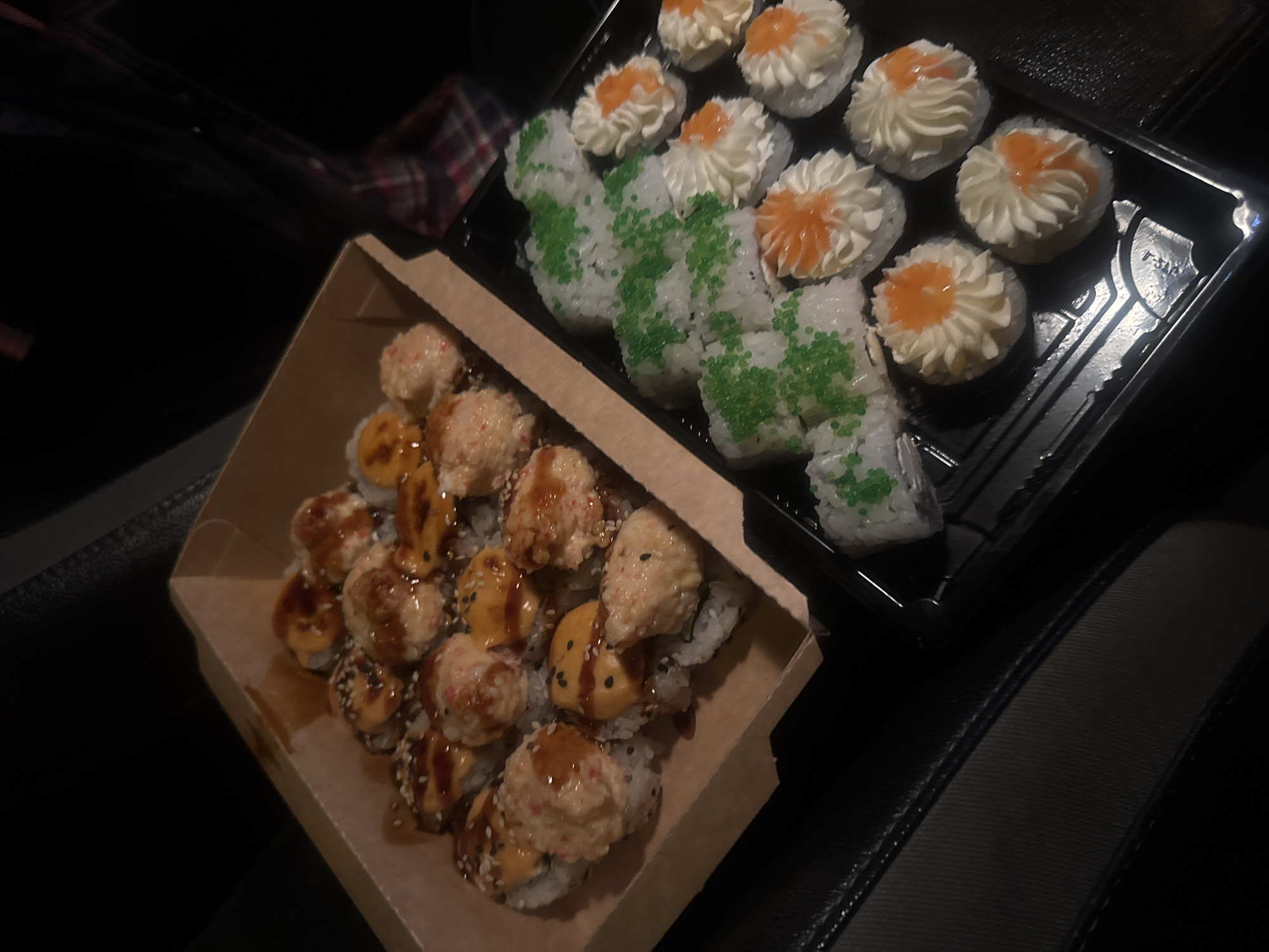 Two containers of sushi are on a dark surface. The container on the left is brown and contains sushi rolls with toppings and sauces. The container on the right is black and contains sushi rolls with white and green toppings. The location may be in Tbilisi, Georgia but there is insufficient evidence to support the claim.