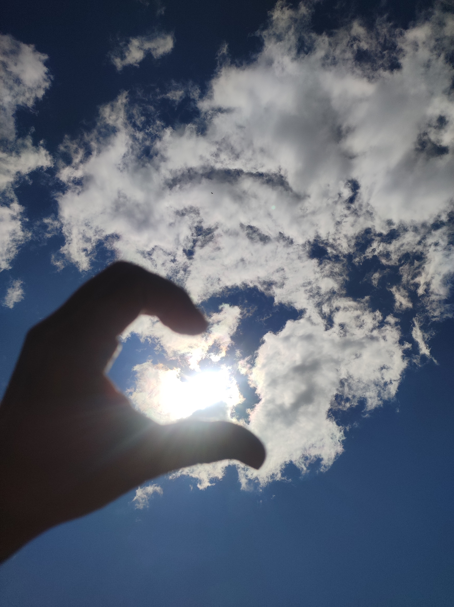 A low-angle, outdoor, close-up shot captures a silhouette of a human hand forming a heart shape against a bright sky. The sun is directly behind the hand, creating a lens flare and obscuring finer details of the hand's structure, though its outline is clearly visible against the sky. The sky itself is a vibrant blue, partially filled with scattered white clouds. The sun's rays are visible, radiating outwards and through the clouds. There is no visible indication of the city of Фоки, Russia, in this image.