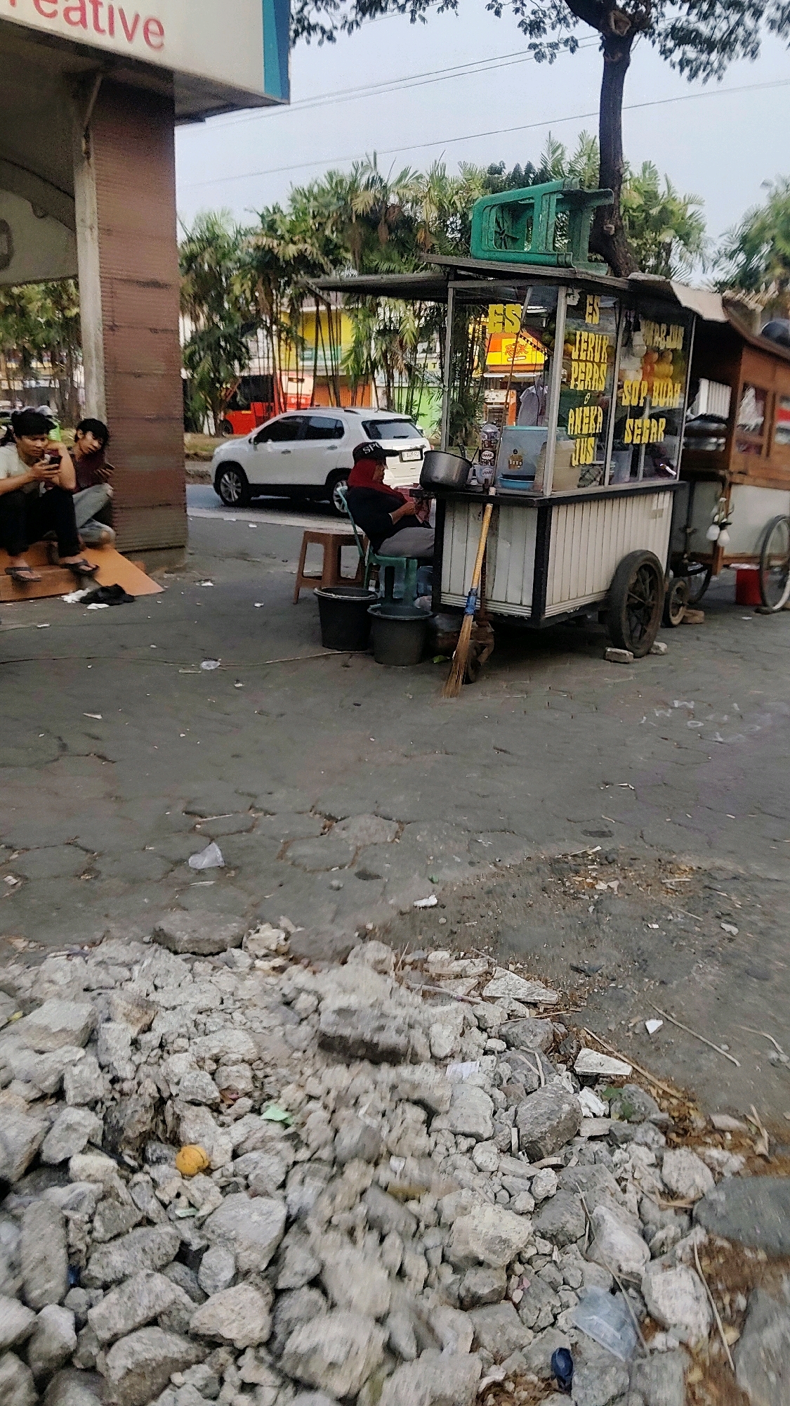 The image captures a street scene in Mekarmukti, Indonesia, focusing on a beverage cart. A white SUV is parked on the asphalt road in front of a building with palm trees visible behind it. To the left, three individuals are seated, with two of them looking at their phones. The beverage cart, a mobile unit on wheels, is positioned on the roadside. A person wearing a red head covering and a black shirt sits on a green stool beside the cart, appearing to be tending to it. The cart itself is adorned with signage advertising various drinks, including "ES JERUK PERAS" (squeezed orange juice) and "SOP BUAH SEGAR" (fresh fruit soup). A broom leans against the cart, and several buckets are placed nearby. In the foreground, a pile of rubble, consisting of gray concrete and stone debris, is scattered across the cracked pavement. The environment suggests an urban setting with signs of construction or demolition.