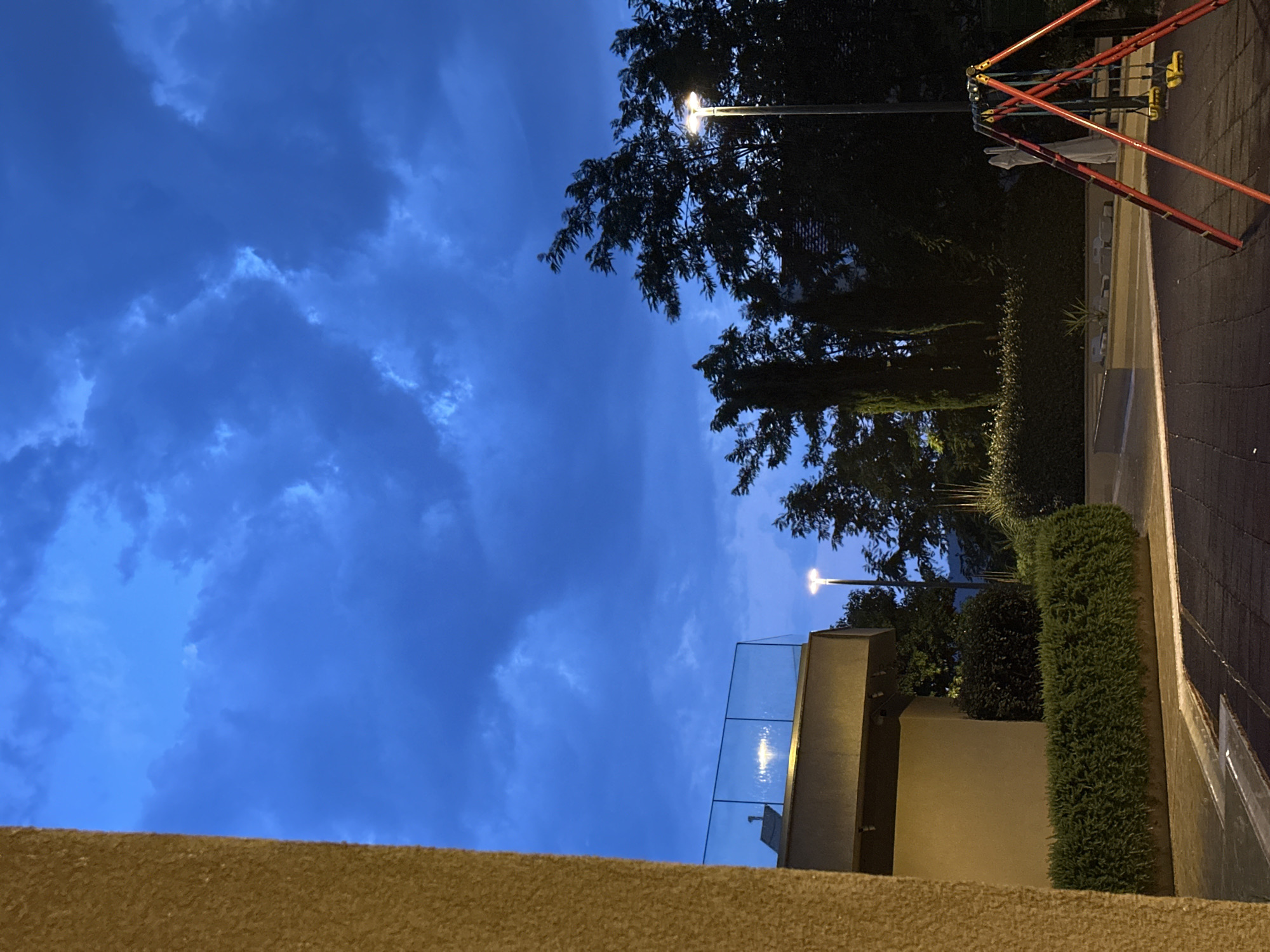 The image captures an outdoor scene at dusk or early evening. The sky is a deep, rich blue, filled with dramatic, dark clouds that suggest an approaching storm or recent rainfall. The lighting indicates it's twilight, with artificial lights beginning to illuminate the scene.
In the foreground, a textured, light brown wall runs horizontally across the bottom of the frame, suggesting a building or balcony. Above this wall, a modern structure is visible with a glass railing that reflects the artificial lights, creating a shimmering effect. Beyond this structure, dense, dark green bushes and trees form a natural barrier.
To the right of the frame, a paved area, possibly a walkway or court, is visible. It has a dark, textured surface with faint lighter lines, suggesting it is made of asphalt or a similar material. A red and yellow metal structure, possibly part of a playground or recreational equipment, stands against the backdrop of trees and the twilight sky.
Two streetlights are visible. One is in the middle ground, its light casting a glow on the glass railing and surrounding foliage. Another, further in the background, is a single point of light against the dark sky, partially obscured by trees.
The scene appears to be in a residential or public outdoor area, likely a park or a residential complex, given the presence of playground equipment and landscaped greenery. The overall mood is one of tranquil transition from day to night, with a hint of atmospheric drama from the cloudy sky. No people are visible in the frame. There is no visible text in the image.