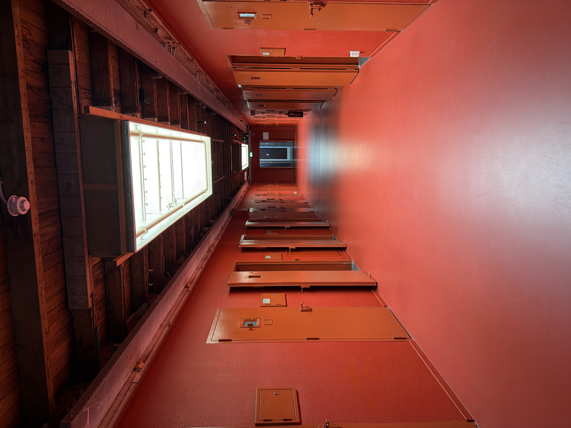 The image depicts a long, narrow hallway with bright red walls and floor, illuminated by a large skylight in the ceiling and fluorescent lights. The hallway is lined with numerous identical orange doors, some of which are ajar, suggesting individual cells or rooms. These doors are fitted with small square windows and metal latches. On the left wall, there are also small square access panels with circular locks. The ceiling is made of exposed wooden beams with visible conduits and fixtures. The overall aesthetic is stark and institutional, reminiscent of a prison or a similar facility. The scene is devoid of people and any visible signs of activity.
This hallway is located in the former **Bijlmerbajes prison in Amsterdam**. While the prompt mentions The Hague, this specific image is of a facility in Amsterdam.