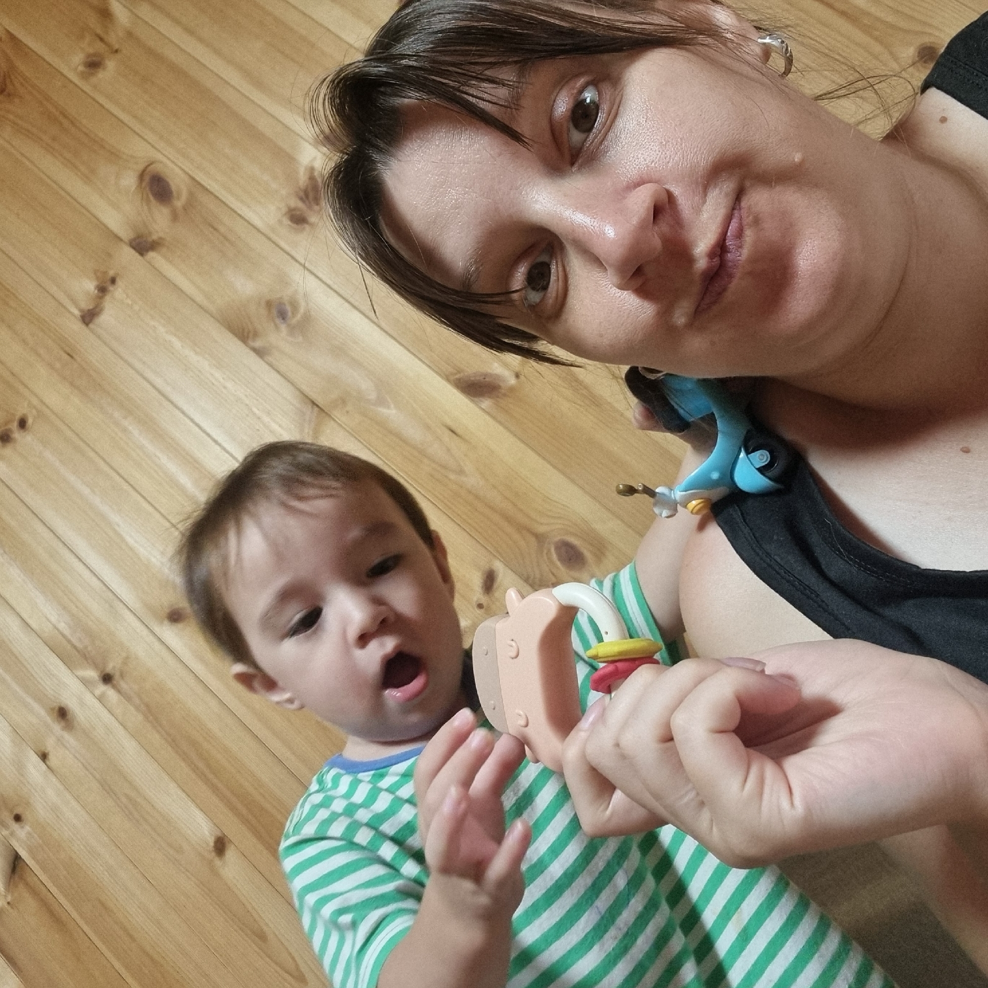A close-up selfie shows a woman and a child against a wooden paneled background in Dunnae-myeon, South Korea. The woman, in the foreground, is wearing a black tank top and silver hoop earrings. Her face is turned towards the camera, with wide eyes and a slightly pursed mouth. A small, blue toy bird is perched on her shoulder. In the background, to the left, a young child wearing a green and white striped t-shirt looks at a pastel-colored toy rattle. The child's mouth is open, as if in mid-speech or surprise. The wooden paneling suggests an indoor setting, possibly a home environment.