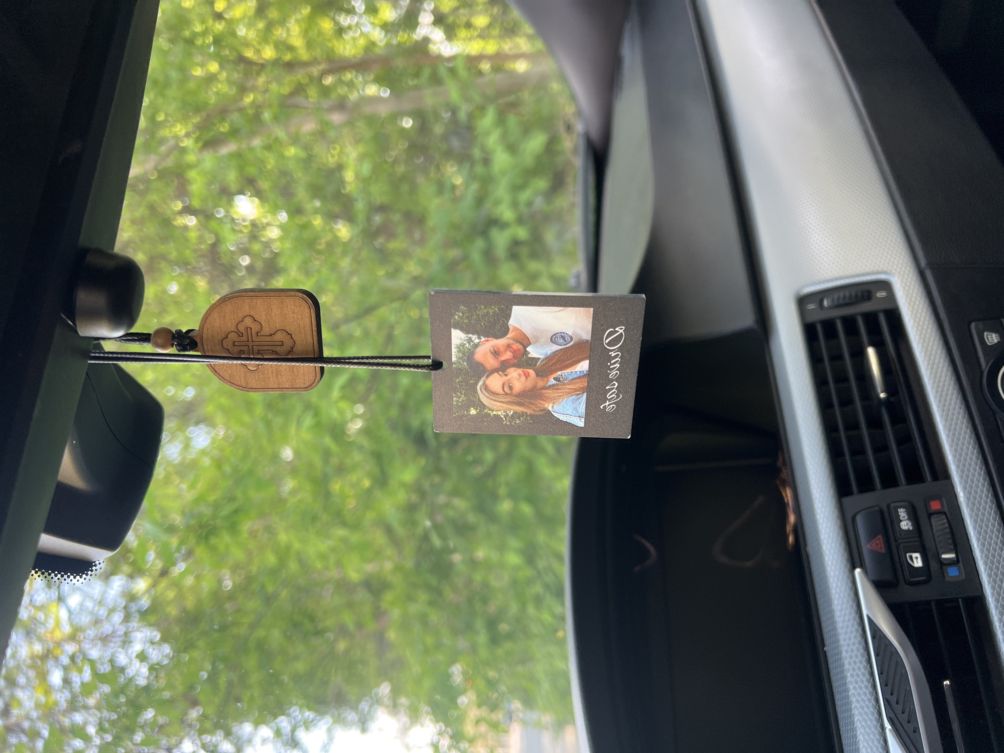 The image presents a view from inside a car, looking forward through the windshield. In the foreground, hanging from what appears to be the rearview mirror (partially visible at the top left), are two decorative items.
On the left, a wooden ornament shaped like a small shield or tablet features a carved cross design, possibly an Orthodox cross, attached by a black string with a small wooden bead. To its right, a small rectangular photo print, also suspended by a string, displays a cheerful young couple. The man has short dark hair and wears a white polo shirt, while the woman, with long blonde hair, wears a denim jacket. Both are smiling. Vertically written along the right edge of the photo is the word "Divine."
The interior of the car is visible on the right side of the frame, showcasing a modern dashboard with a textured silver trim contrasting with black plastic elements. A car air vent is clearly seen, along with a set of control buttons, including a prominent red triangular hazard light button and a door lock/unlock button.
Through the windshield, the background shows lush green foliage, indicating trees or bushes, softly blurred by a bokeh effect, suggesting natural sunlight. The bright conditions point to daytime. This scene is set within a car in Plovdiv, Bulgaria.