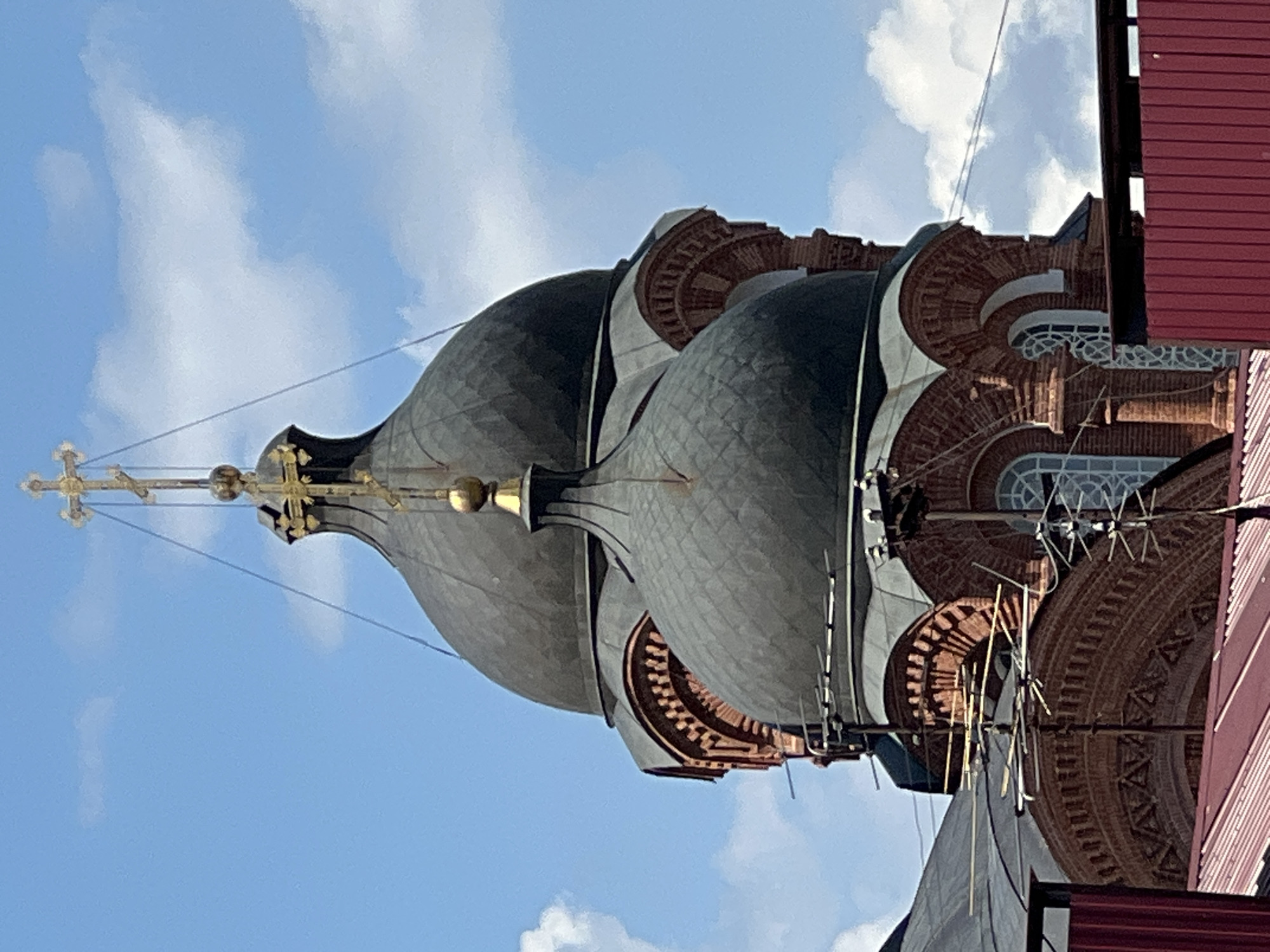 The image shows the domes and facade of a red brick church, likely Orthodox Christian, under a bright blue sky with scattered white clouds. The church features multiple dark, rounded domes, with the largest dome topped by a golden cross. Smaller domes and architectural details, such as arched windows and brickwork patterns, are visible on the structure. In the foreground, a red roof and part of a red fence are visible. Antennas are mounted on the facade of the church, suggesting it is in an urban setting. The style of the architecture suggests it could be located in Krasnodar, Russia, a city known for its numerous Orthodox churches. No people are visible in the image.