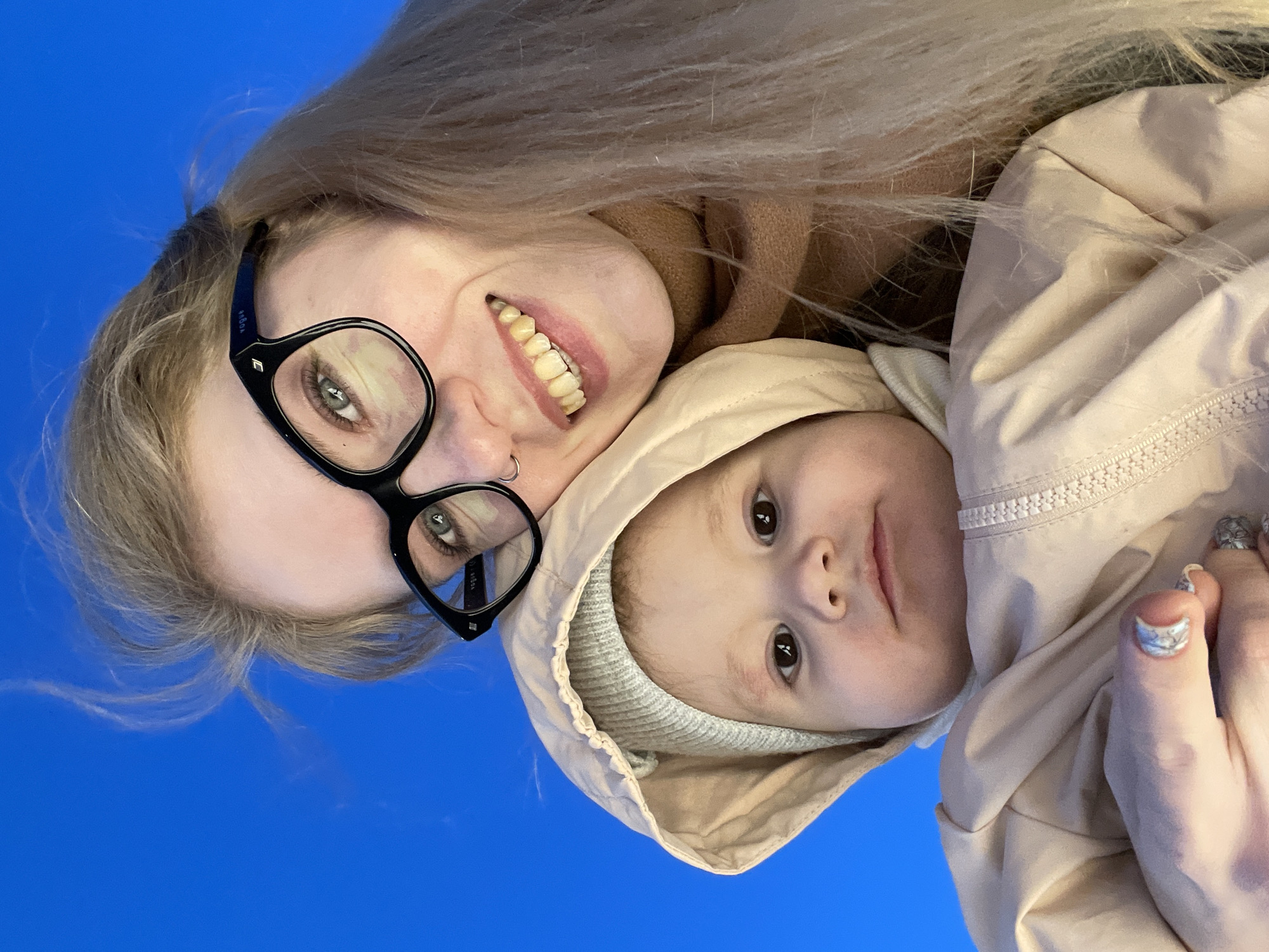 A woman with blonde hair and glasses is holding a baby wrapped in a beige hooded jacket. The woman is smiling and wearing a nose ring. The baby is looking at the camera. The background is a solid blue. The location is not discernible.