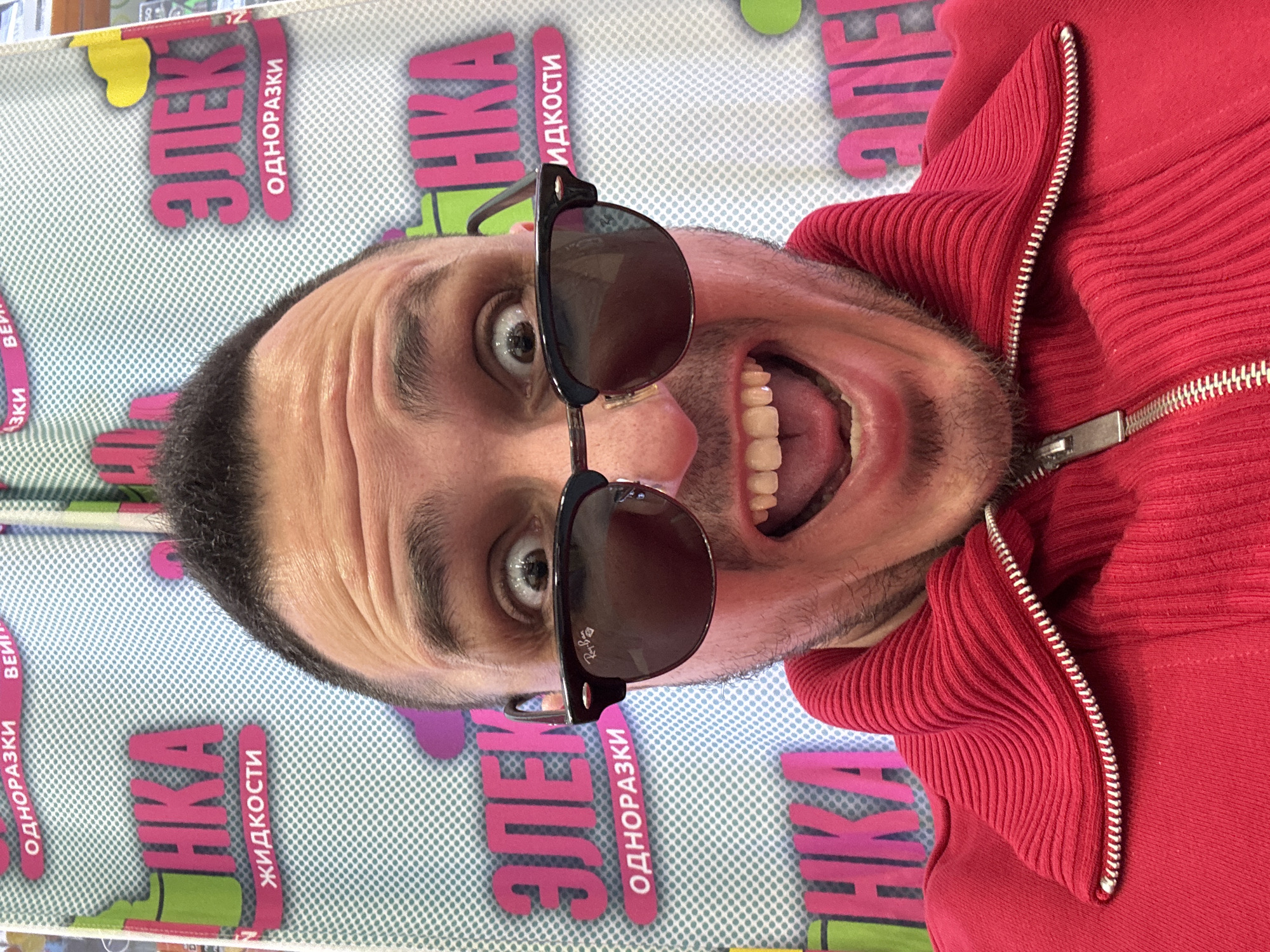 A man with sunglasses poses in Saint Petersburg, Russia. He is wearing a red sweater and grinning widely. The backdrop appears to be an advertising display with the word "ЭЛЕКТРОНИКА" (Electronics) in Cyrillic.