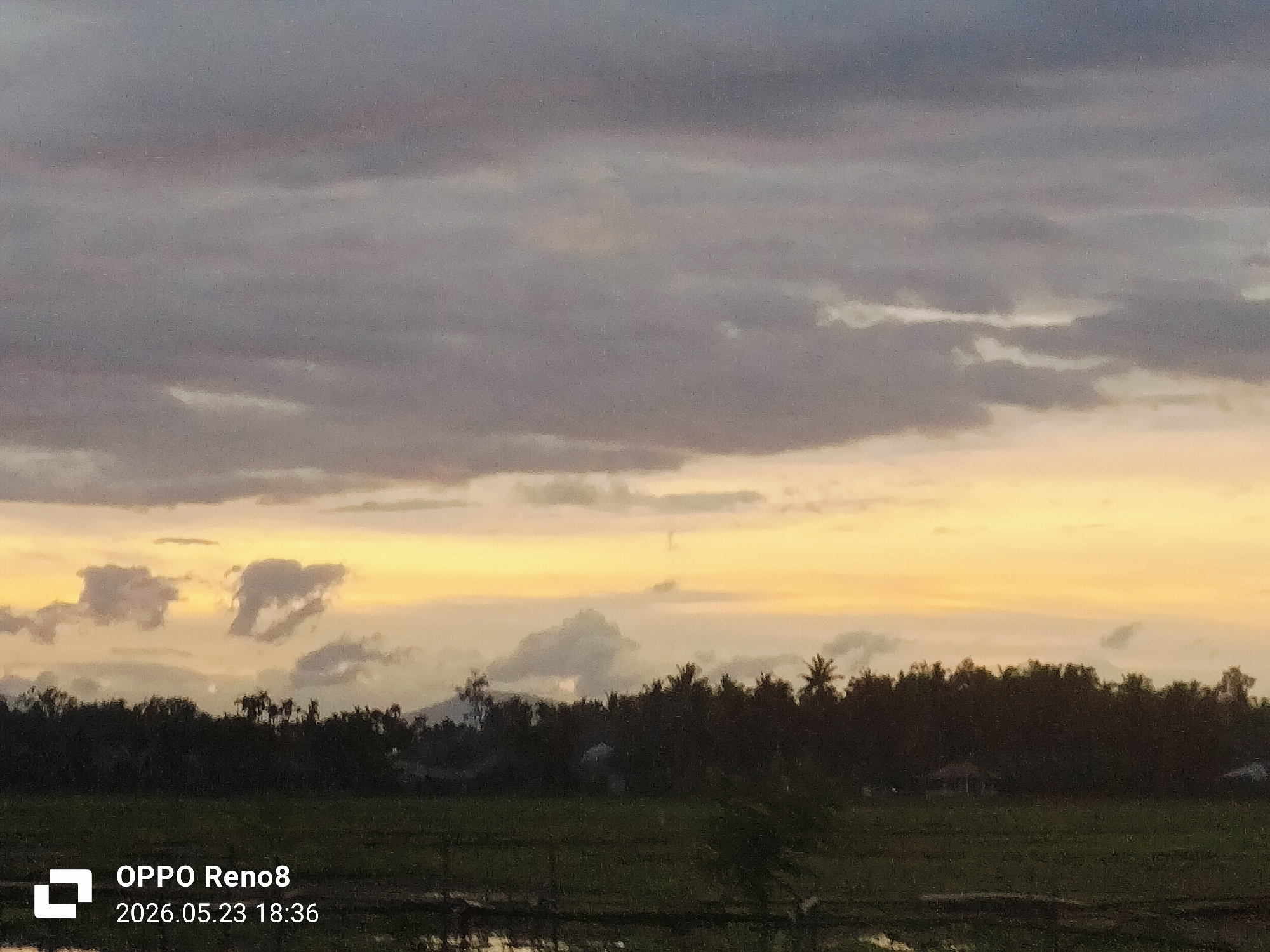 The image depicts a twilight scene in Kampungjumpa, Indonesia. The foreground shows a grassy field. A dark treeline extends across the midground, with silhouettes of various trees, including palm trees, against a vibrant, multicolored sky. The sky transitions from gray and purple at the top to orange and yellow near the horizon. Cloud formations are visible throughout. In the bottom left corner, there is a watermark indicating the image was taken with an OPPO Reno8 on May 23, 2026, at 18:36.