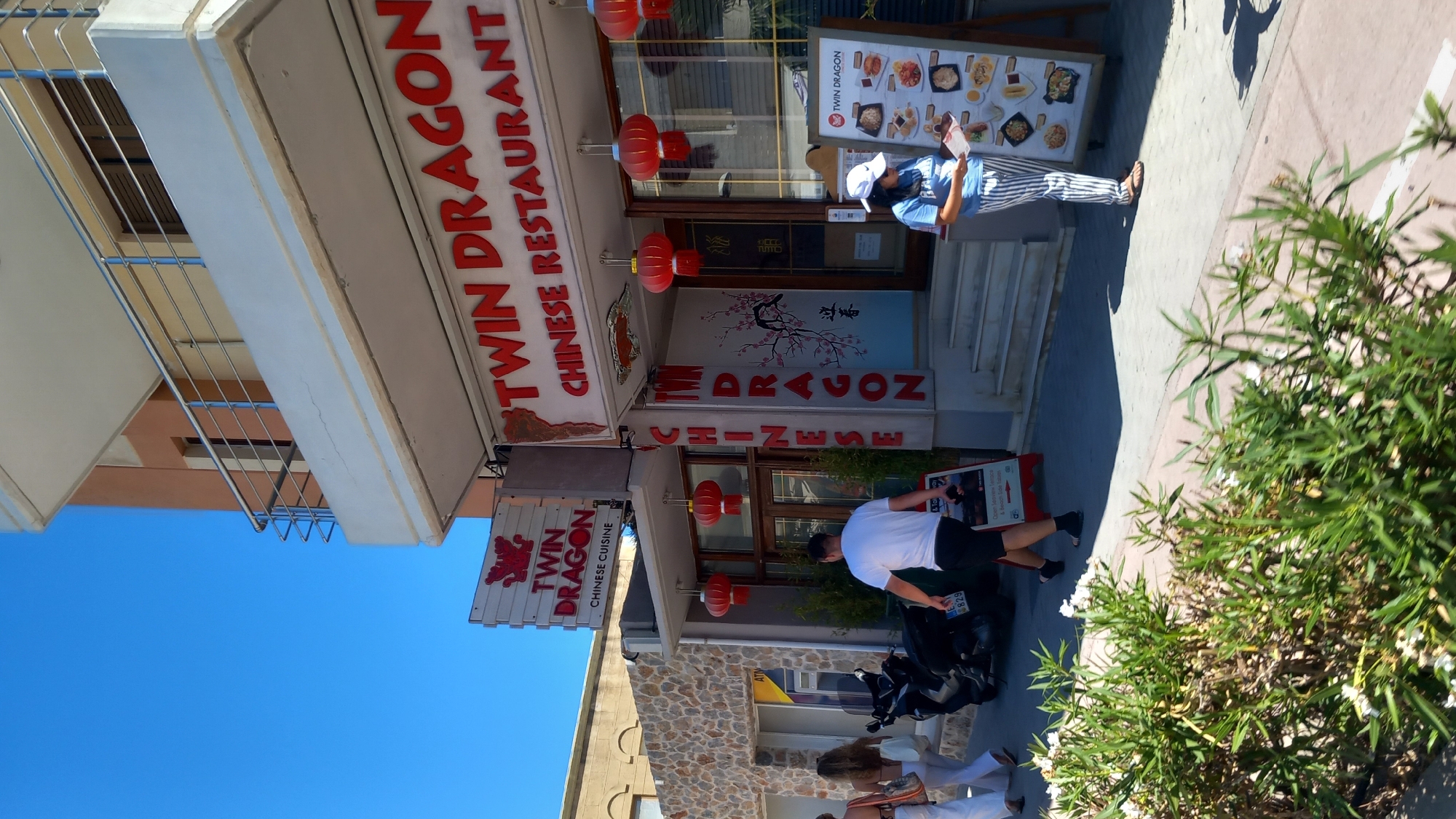 The image depicts the exterior of "Twin Dragon Chinese Restaurant" located on the island of Kos, Greece. The restaurant's facade features prominent red signage with the restaurant's name and "CHINESE CUISINE" in English. Red Chinese lanterns are hung outside the entrance. A menu board displaying various dishes is positioned on the sidewalk in front of the restaurant.
Several people are visible. Two women in white clothing are walking away from the camera towards the left side of the frame. A man in a white t-shirt and black shorts is walking away from the camera towards the right, approaching the restaurant entrance. A woman wearing a blue t-shirt and striped pants is standing in front of the menu board, examining a document.
A black motorcycle is parked on the sidewalk to the left of the restaurant. Green bushes with white flowers are in the foreground, partially obscuring the sidewalk and street. The sky is a clear blue. The architecture suggests a Mediterranean coastal town setting.