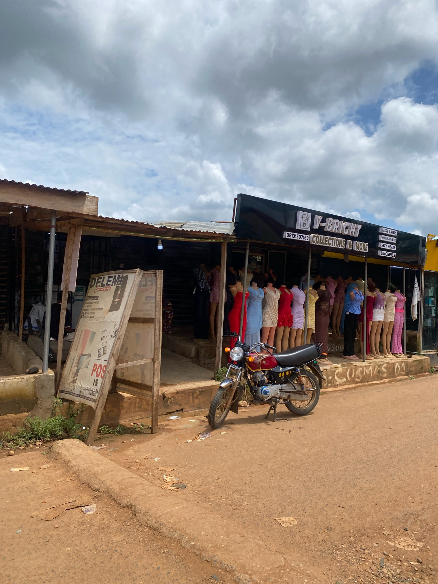 The image depicts a street scene in Iworoko, Nigeria, featuring a shopfront with mannequins displayed outside. The shop, named "V-BRIGHT COLLECTIONS & MORE", has a black awning with white lettering and lists various items for sale, including "BAGS & SLIPPERS", "WHOLESALE SALES & RENTALS", "SEXY WEARS", and "LINGERIES". A row of mannequins, dressed in colorful dresses, are lined up in front of the shop.
To the left of the "V-BRIGHT" shop, there is another structure with a corrugated iron roof and wooden supports. A large, leaning sign for "DELEMI CONCEPTS" is positioned in front of this structure. This sign advertises "CHANGING OF PHONES", "REPAIR", "SALES OF PHONES & ACCESSORIES", and "POS IS AVAILABLE HERE".
A red and silver motorcycle is parked in front of the "V-BRIGHT" shop. The ground is a dirt road, with some debris scattered around. The sky above is partly cloudy. The overall impression is of a bustling market area with commercial activities taking place.