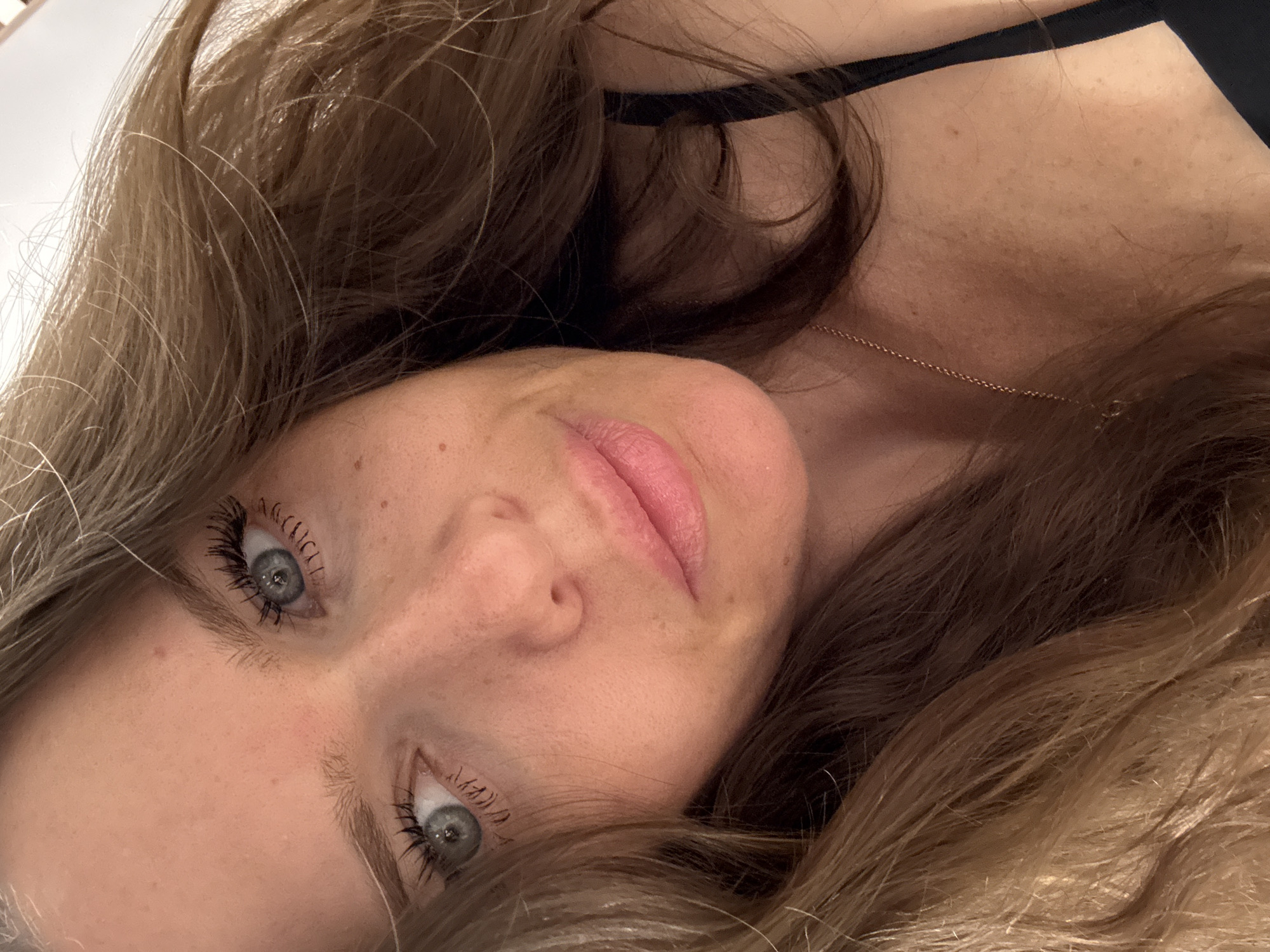 The image shows a close-up, overhead shot of a woman's face and upper chest. She has long, wavy brown hair with lighter blonde highlights, framing her face. Her eyes are blue-grey and her lips are lightly colored. She is wearing a black top, and a thin gold necklace is visible around her neck. The background appears to be a plain, indoor setting, possibly a room. There is no indication of the city of Moscow, Russia, or any specific location within it.