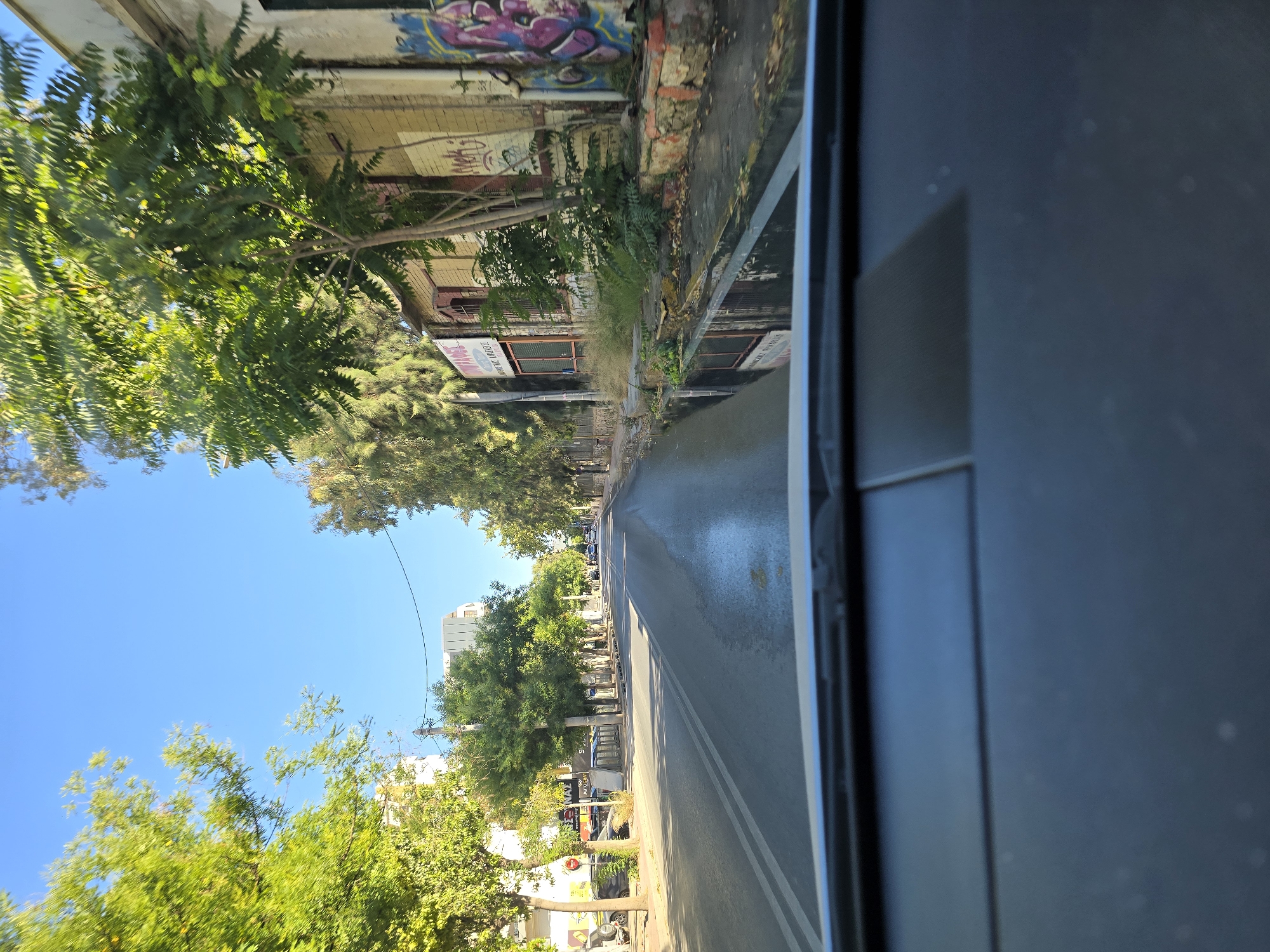 The image depicts a street scene in Athens, Greece, viewed from the perspective of inside a vehicle. The road is asphalt with white lane markings and appears wet, reflecting the clear blue sky and surrounding trees. Lush green trees line both sides of the street, casting shadows. Buildings are visible along the right side of the road; one has graffiti on its exterior. There are several parked cars and a motorcycle on the left side of the street. In the distance, more trees and buildings can be seen, with a modern structure in the background. The dashboard of the vehicle is in the foreground, slightly out of focus. The overall impression is of a bright, sunny day in a moderately urban environment.