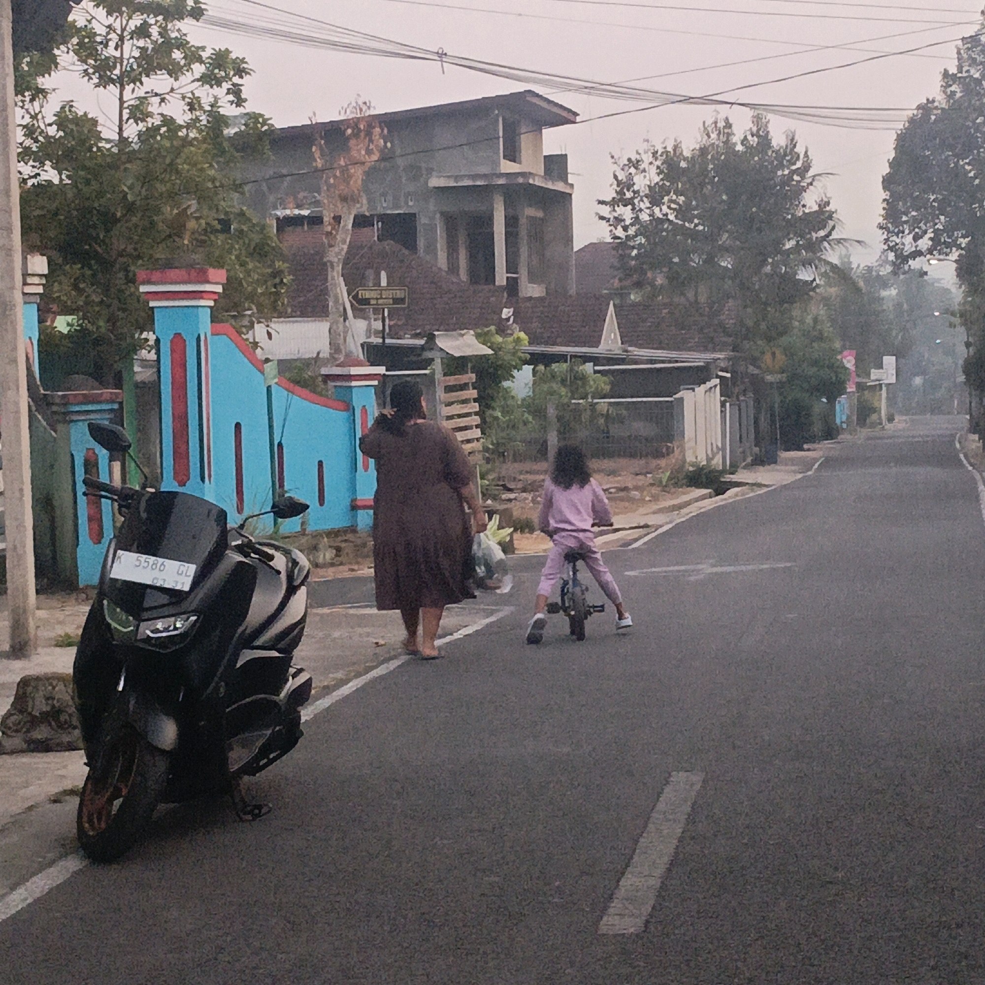 In the city of Salatiga, Indonesia, a street scene captures a mother and child during daylight hours. The mother, dressed in a dark brown abaya, walks along the right side of the asphalt road, carrying a plastic bag. Beside her, a young girl in a light pink tracksuit rides a small bicycle. A black motorcycle is parked on the left side of the road, its license plate visible. The background features a blue and red painted wall, a partially constructed building, trees, and utility lines overhead. The atmosphere suggests a modest urban or suburban setting.