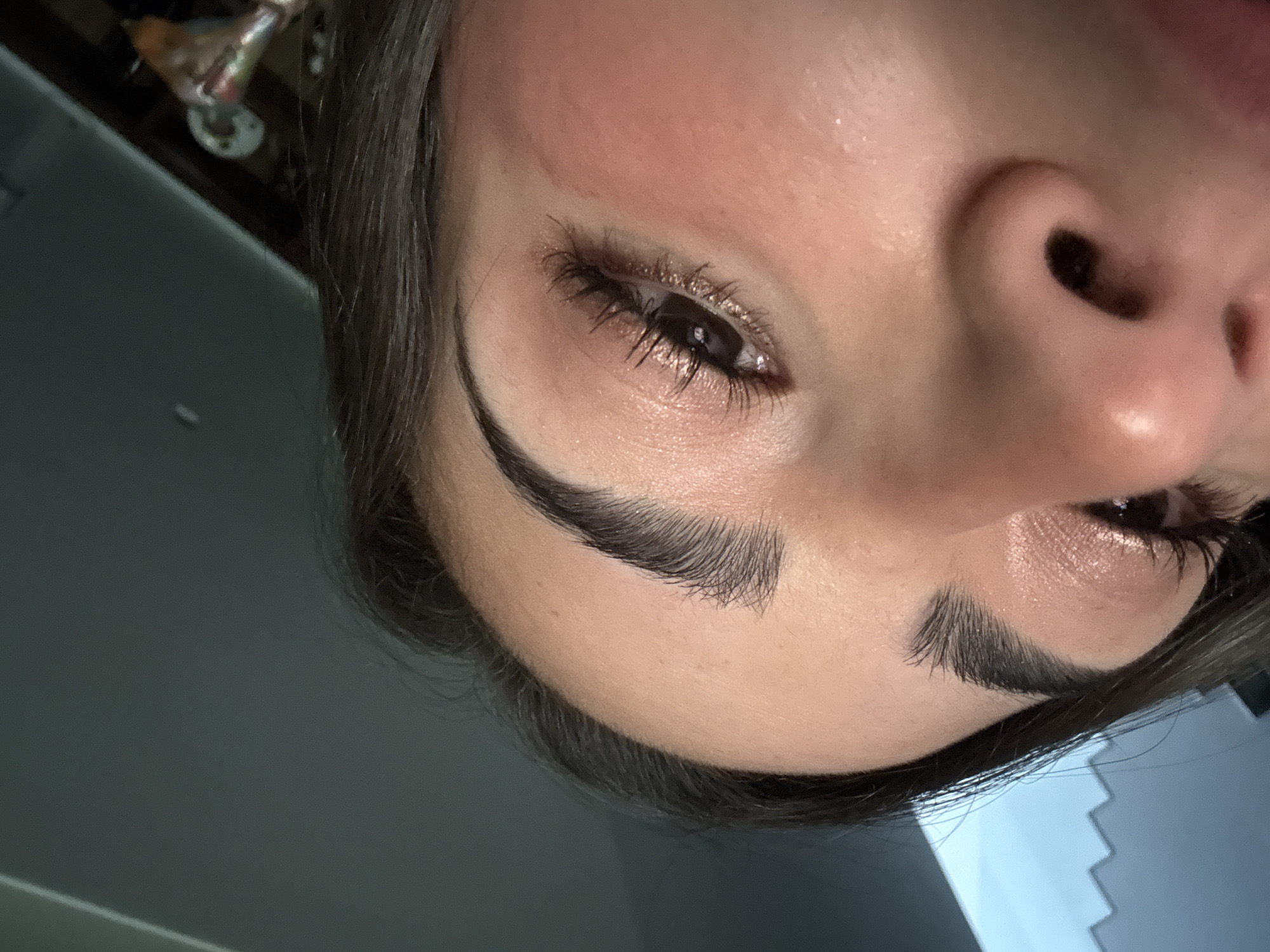 The image displays a close-up, angled view of the upper right quadrant of a person's face, located in San Bernardino Tlaxcalancingo, Mexico. The subject's right eye is the focal point, featuring prominent dark eyelashes, consistent with mascara or lash extensions, and shimmery, light-toned eyeshadow on the eyelid. The iris of the eye is dark brown. The right eyebrow is dark, thick, and well-defined. The skin, of a fair to medium tone, exhibits a slight sheen, with a reddish blush visible on the upper cheek below the eye. Dark hair frames the forehead and temple area on the left side of the frame. The bridge of the nose and a portion of the right nostril are visible in the lower right, indicating an upward camera angle. The background in the upper left is a flat, dark teal-gray surface with a diagonal light-colored line, while the upper right background contains indistinct, darker shapes, including vague metallic elements suggesting indoor fixtures or furniture. The perspective is from a low, upward angle, directed towards the subject's face.