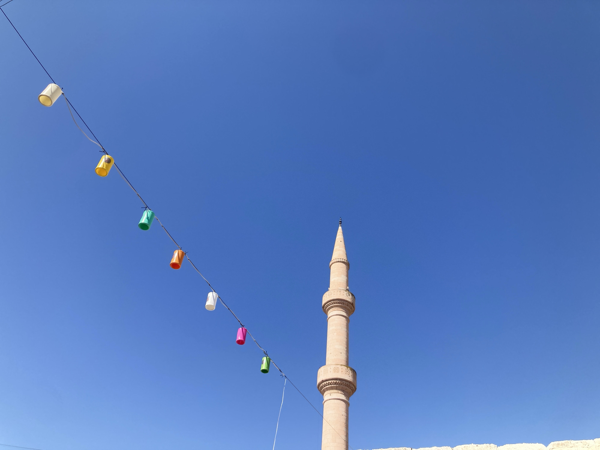 A string of colorful cylindrical lanterns hangs across a clear blue sky, with a tall, light-colored minaret of a mosque visible in the background. The lanterns, in shades of white, yellow, green, orange, and pink, are spaced out along a wire that runs diagonally from the left to the right of the frame. The minaret, made of stone, has intricate decorative bands and tapers to a pointed spire. The scene appears to be outdoors, likely in the city of Avanos İlçe Merkezi, Turkey, given the architectural style of the minaret. The bottom of the frame shows a hint of a stone structure, possibly a wall or building. There are no people or animals visible in the image.