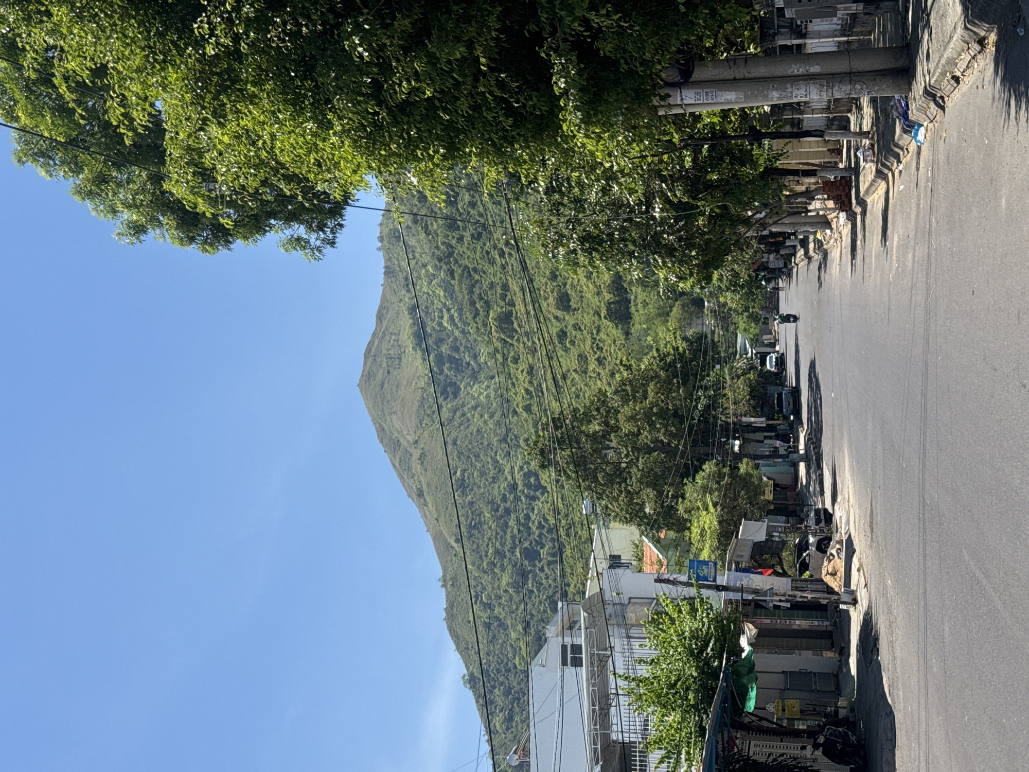 The image depicts a street scene in Bac Nha Trang Ward, Vietnam, with a prominent green mountain range in the background. The street is paved and lined with trees on the right side, casting shadows on the sidewalk. On the left side of the street, there are buildings, some appearing to be shops with awnings, and residential structures. A few vehicles are visible on the street, including a car parked on the side and a motorcycle being ridden. There are also some pedestrians present. Overhead, a network of power lines crisscrosses the sky. The weather appears to be clear and sunny, with a bright blue sky. The overall impression is of a busy, everyday street in a tropical or subtropical urban environment, with a natural landscape forming the backdrop.