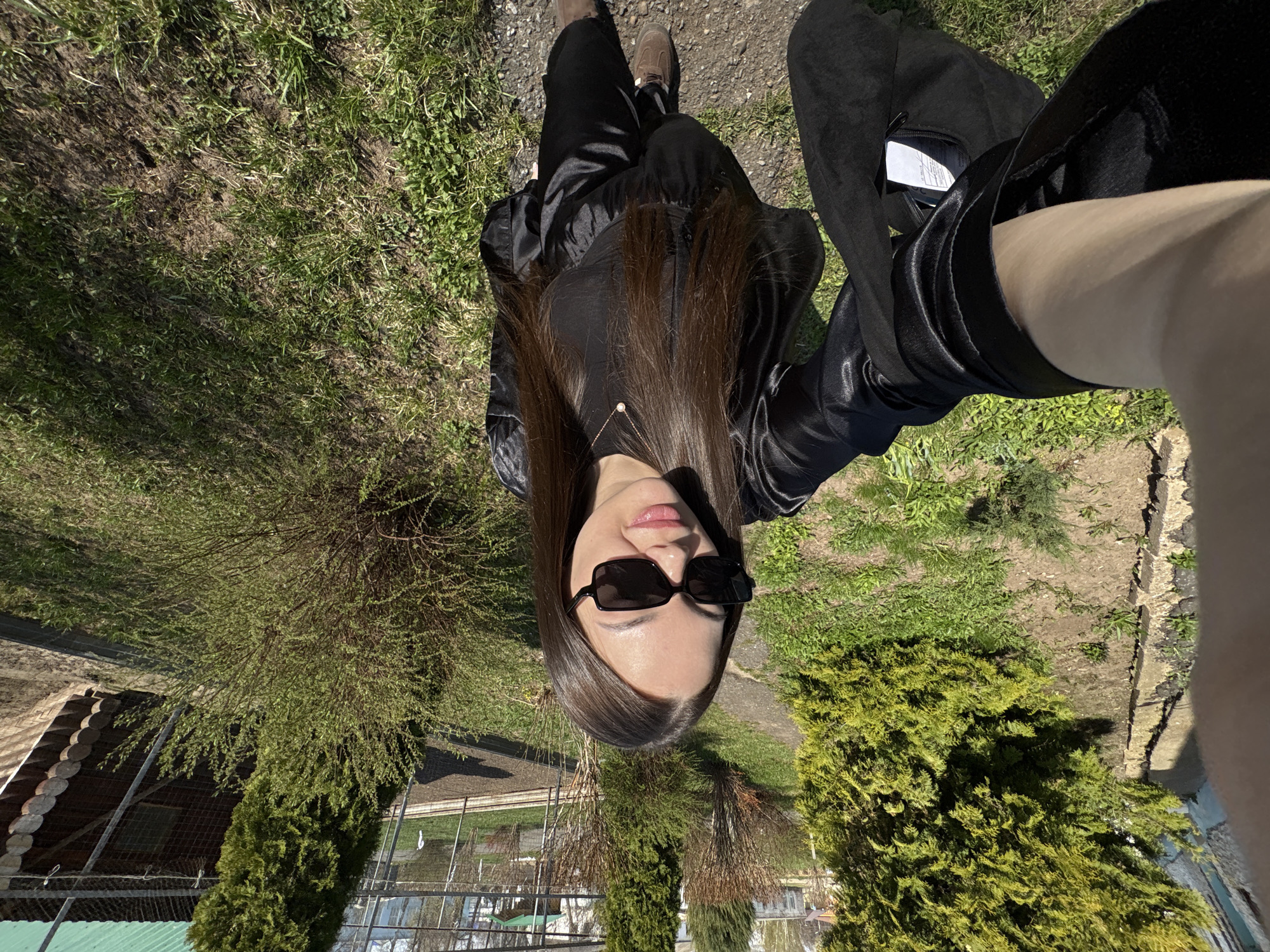 A photograph shows a woman in sunglasses, wearing a black outfit and tan shoes, seated outdoors on a grassy area next to bushes and trees. The location is identified as Chaikovsky, Russia, indicated by the architecture and landscaping. The photo appears to be taken from an upward-looking angle. A building with a brown roof is visible in the background, behind a fence.