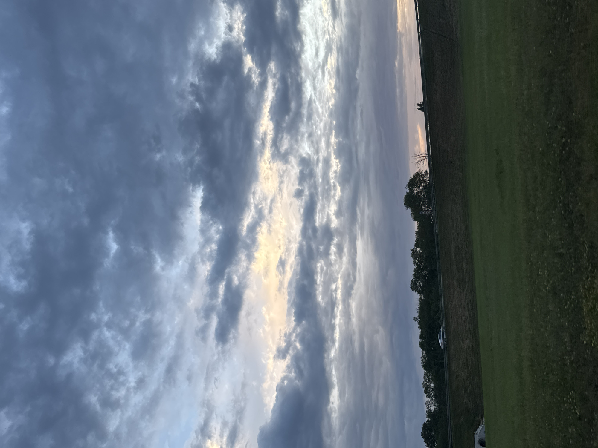 This media file depicts a landscape scene with a focus on the sky and a grassy field. The sky is filled with dramatic clouds, ranging from dark, heavy storm clouds to lighter, more wispy formations. Streaks of sunlight break through the clouds in the upper part of the image, suggesting either sunrise or sunset.
The lower portion of the image shows a well-maintained green field, bordered by a dark line of trees and vegetation. A faint fence or barrier runs along the edge of the field. A single, white vehicle is visible on a dark path or road in the distance, partially obscured by the trees. The overall mood is somewhat somber due to the heavy cloud cover, but the sunlight creates a sense of hope or transition. The scene appears to be in a rural or semi-rural area, possibly a park or a field adjacent to a road. The time of day is likely early morning or late evening, given the lighting conditions and cloud formations. There is no visible text in the image.