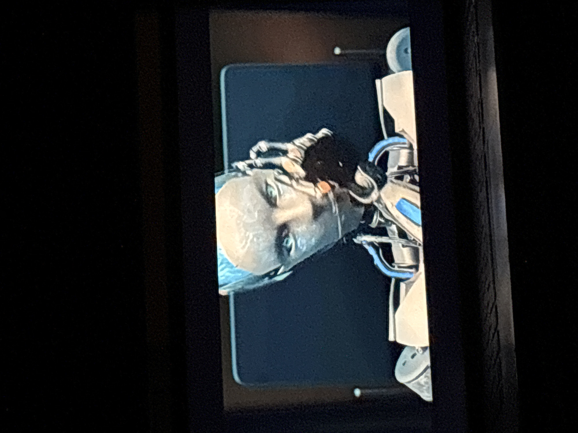 The image presents a close-up of a screen displaying a detailed, human-like robotic or android figure. The subject is primarily the head and upper torso of the robot, rendered with metallic or synthetic textures in light grey and silver tones. The face features realistic eyes with visible irises and pupils, a subtle nose, and mouth area, conveying a neutral and serene expression. Intricate, light blue or teal-colored elements resembling delicate structures or hair are visible around the back and side of the head.
To the robot's left (viewer's right), complex mechanical components are visible, including metallic structures with bright blue accents—possibly tubing or wiring—and white or light grey casing, forming its neck and shoulder. A partial view of a metallic, multi-fingered hand-like structure is also discernible near the head.
The robotic figure is showcased against a dark blue or dark grey screen background with gently rounded corners. This screen is enveloped by a wider, dark, and subtly textured frame, suggesting it's part of a sophisticated display unit or console. The area surrounding this frame is predominantly black. The scene captures a static image, likely a digital rendering or a photograph of a physical model, highlighting advanced technology. The lighting emphasizes the metallic surfaces, creating a sense of depth. There is no visible text, and no specific location, time of day, or weather cues are evident within the image.