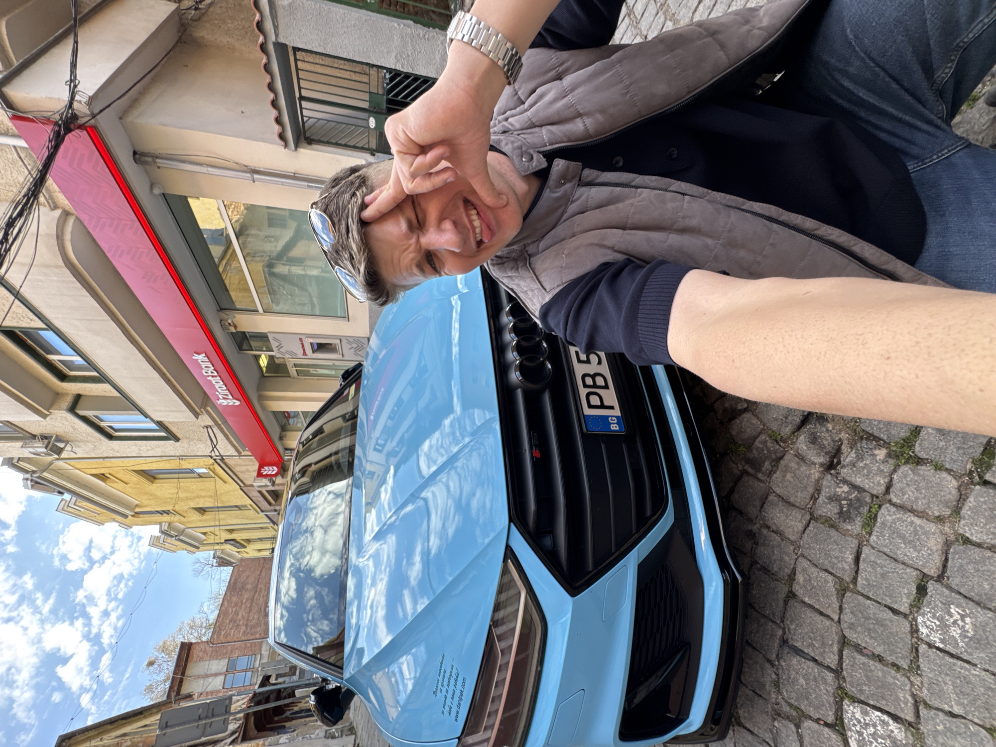 A man is taking a selfie with a light blue Audi car on a cobblestone street. The car has a Bulgarian license plate "PB 5". The man is wearing a vest and jeans. Behind him is a building with a red sign that says "UniCredit Bulbank". The location is likely Plovdiv, Bulgaria, given the presence of a Bulgarian license plate and the bank name.