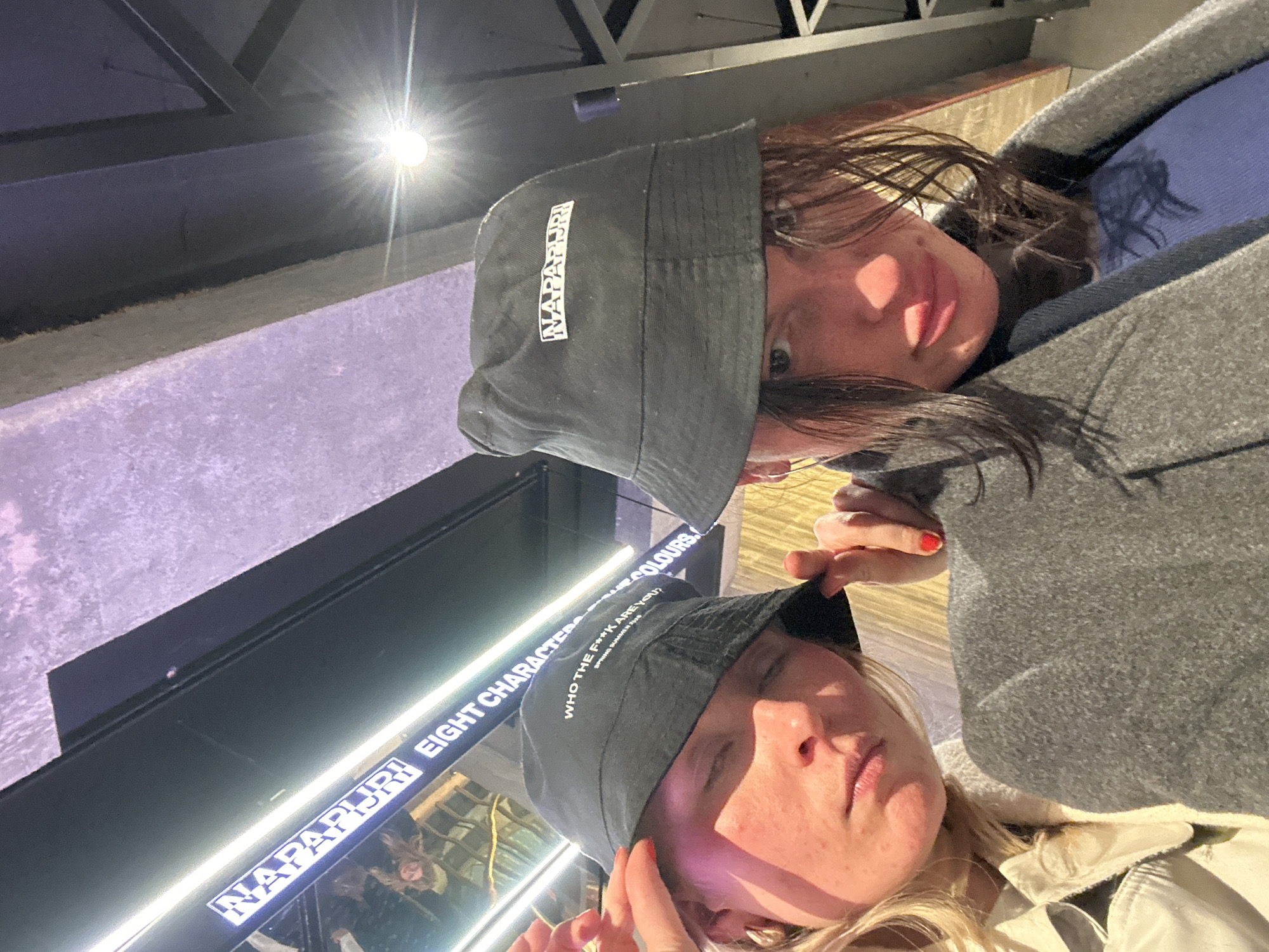 Two women are posing for a selfie. Both are wearing Napapijri brand bucket hats. The woman on the upper right has darker hair and is wearing a grey coat. The woman on the lower left has blonde hair and is wearing a light-colored jacket. Reflected in a window behind them are the words "NAPAPIJRI EIGHT CHARACTERS WHO THE F**K ARE YOU? SPRING SUMMER 2023". The location is a Napapijri store in London, United Kingdom.