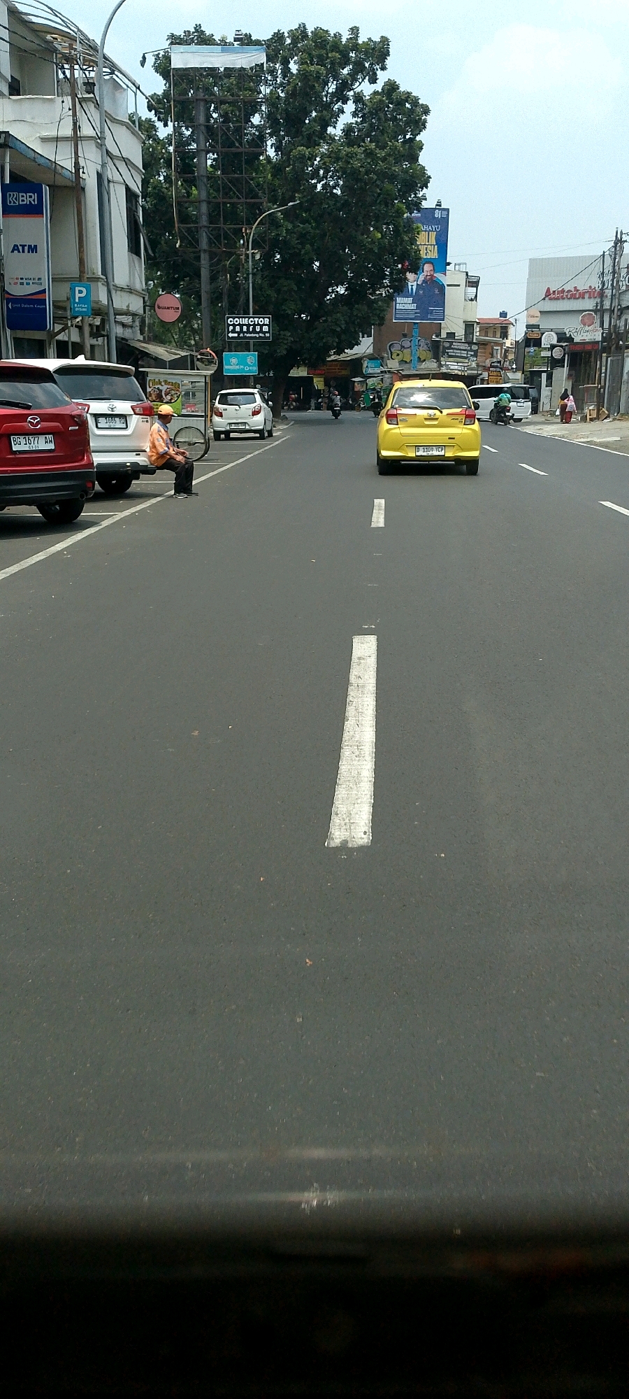 The image depicts a street scene in Bandung City, Indonesia, likely on a cloudy day. In the foreground, a black car's dashboard is visible, suggesting the photo was taken from inside a vehicle. The street itself is asphalt with white lane markings. On the right side of the road, a yellow hatchback car is driving away from the camera, with license plate "D 1159 ICP". Further down the road, other cars and motorcycles are present, and pedestrians can be seen on the sidewalks.
The left side of the street features buildings with various signs. Notably, a "BRI" (Bank Rakyat Indonesia) sign is visible, along with an "ATM" sign, indicating a bank branch. A blue parking sign with a "P" symbol is also present. A large green tree dominates the central background, with a tall, dark metal structure possibly supporting advertising billboards behind it. One billboard shows an image of two men and text in Indonesian.
In the middle ground, on the left side of the street, a man wearing an orange cap and an orange shirt is sitting on what appears to be a cart. He is facing away from the camera and appears to be resting or waiting. Another white car is parked next to him, and a food cart is situated nearby. In the distance, more buildings and businesses are visible, including one with a sign that reads "Autobrite". The overall impression is that of a busy urban street with commercial activity.