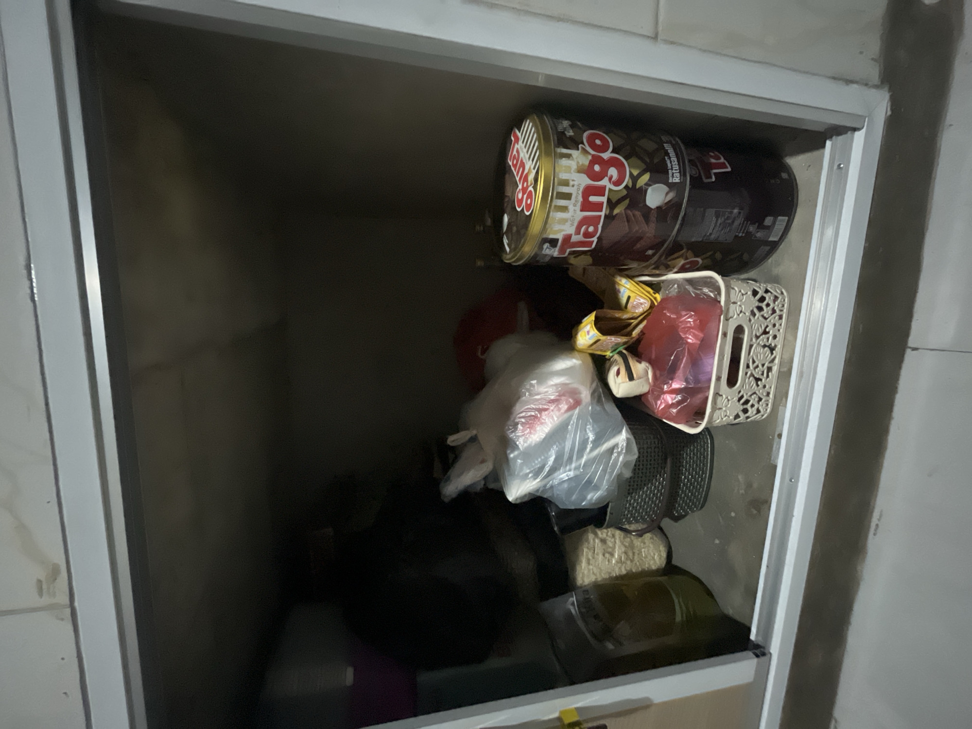 In Kebumen, Indonesia, a storage cabinet's interior is visible, filled with various items. Dominating the right side is a large, round, dark brown tin of Tango wafer snacks. To its left, a white woven basket holds red-wrapped items and yellow snack packages. Behind these, a clear plastic bag contains more items, and next to it is a dark grey woven basket. A large, clear plastic container filled with pale yellow granular material sits on the left side, beside a dark green object. The interior of the cabinet is dimly lit, with a neutral-colored ceiling above.