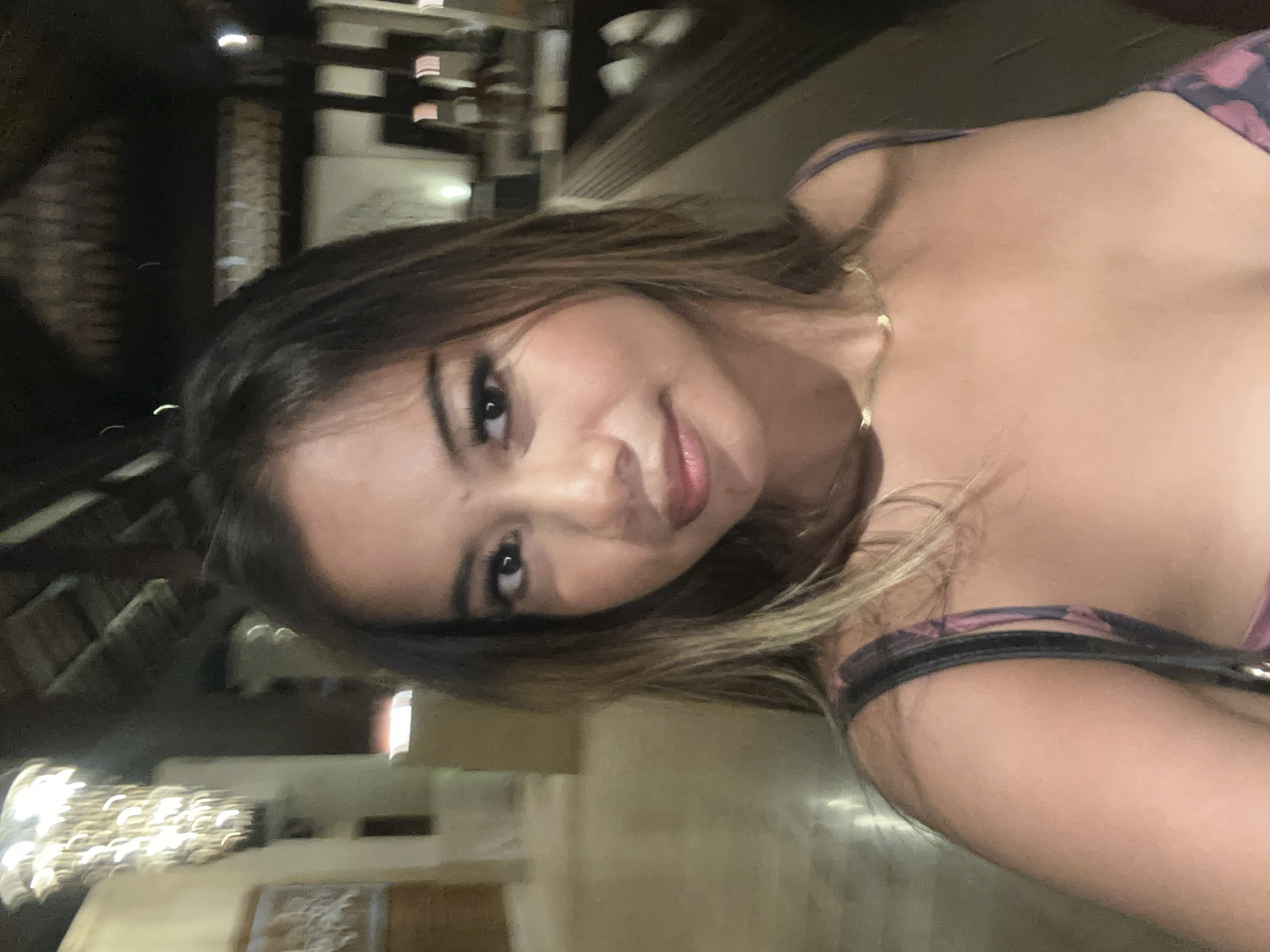 The image features a close-up selfie of a young woman, captured indoors, likely during the evening or night due to the prominent artificial lighting and dark ambiance. She is positioned diagonally in the frame, looking directly at the camera with a calm, slightly smiling expression.
The woman has light brown hair with blonde highlights, styled loosely around her face. Her facial features are clear, showcasing her eyes, nose, and lips. She is wearing a strappy top or dress with a dark pattern, possibly incorporating shades of purple or pink. A delicate gold chain necklace is visible around her neck. Part of her shoulder and arm are also visible in the lower right portion of the frame.
The background is softly blurred, indicating an indoor setting such as a restaurant, bar, or hotel lobby. Distinguishable elements include dark architectural details like beams, possibly some textured wall surfaces, and various light sources. In the lower left, a chandelier-like lighting fixture with multiple glowing elements is visible, contributing to the warm, dim lighting of the scene. Other blurred lights can be seen scattered in the background.
The activity is a self-portrait, or selfie, with the woman posing for the camera. Given the provided location context, this scene is set in Kuta Selatan, Indonesia. There is no visible text in the image.