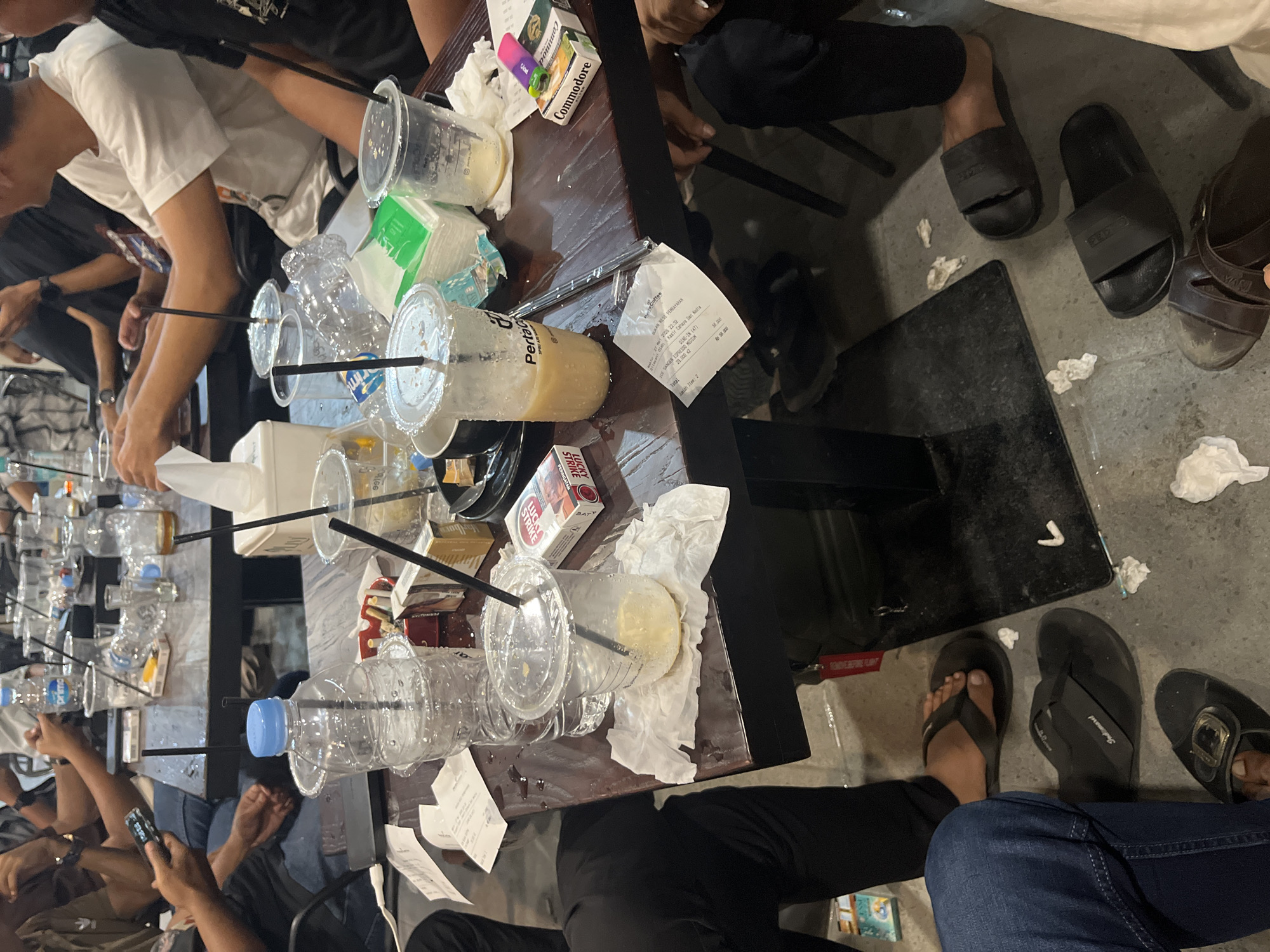 The image depicts a cluttered table in what appears to be a cafe or similar establishment in Meunasahceubrek, Indonesia, as evidenced by the "Pertamina Coffee" receipt. The table is covered with numerous empty plastic cups, some with straws, and empty water bottles. There are also cigarette packs on the table, including brands like "Commodore" and "Lucky Strike". Pieces of crumpled tissue paper are scattered around. People are seated around the table, partially visible in the image. Some individuals are using their phones. The floor around the table is concrete and has discarded tissue and debris. There are various types of footwear visible, including flip-flops and sandals.