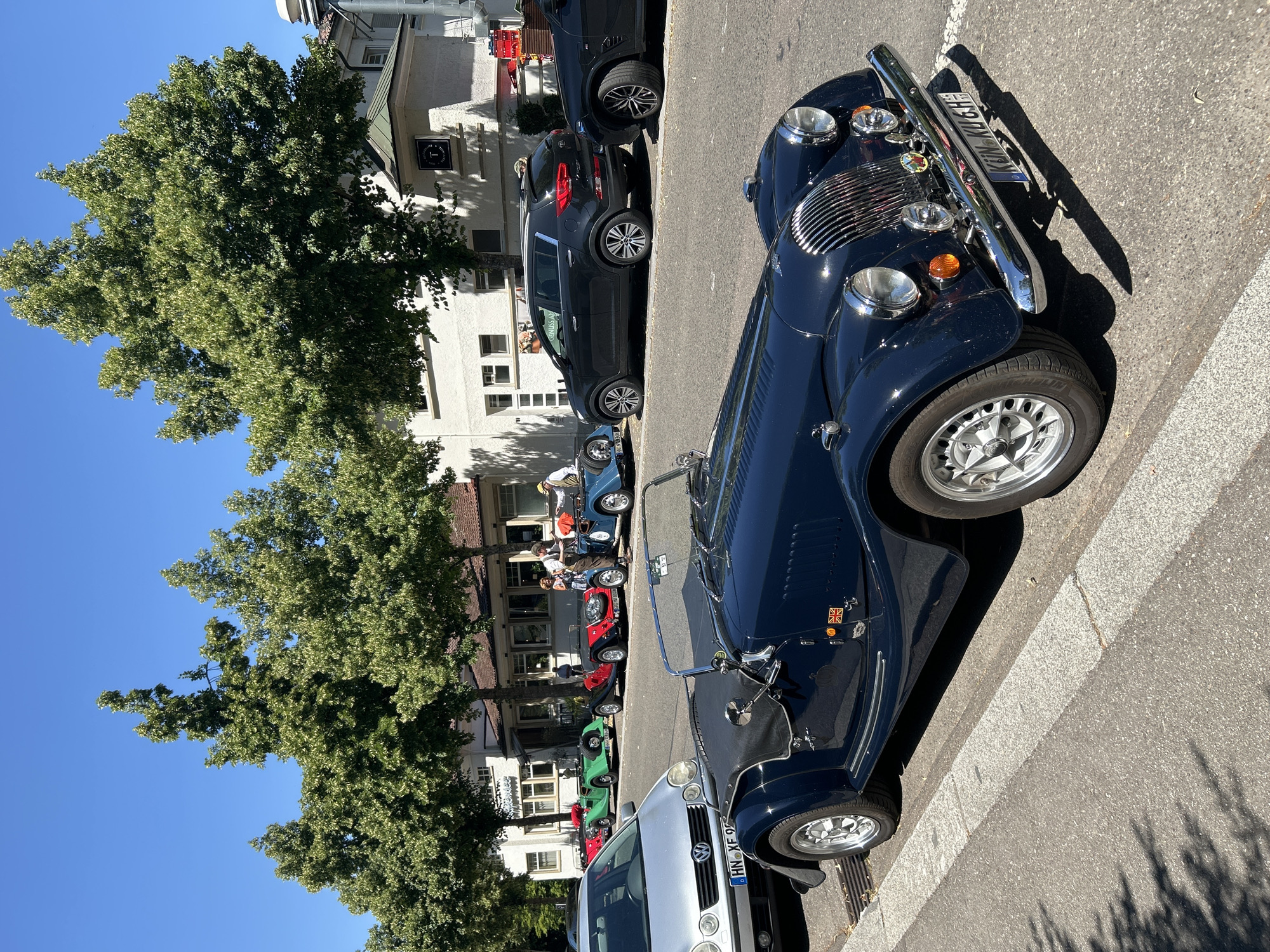 A dark blue convertible classic car is parked on the right side of a street in Heilbronn, Germany. The car is in the foreground, with its front end visible. It has a chrome grille, round headlights, and a license plate that reads "KUN-KU 6H". Other cars are parked behind it, including a silver Volkswagen Polo, a green classic car, a red and black classic car, and a blue convertible classic car. A dark grey SUV is also visible further down the street. Several trees with green foliage line the street, and there are buildings in the background. A few people are standing and walking on the sidewalk. The sky is clear and blue. The scene appears to be during the day, with sunlight casting shadows on the street.