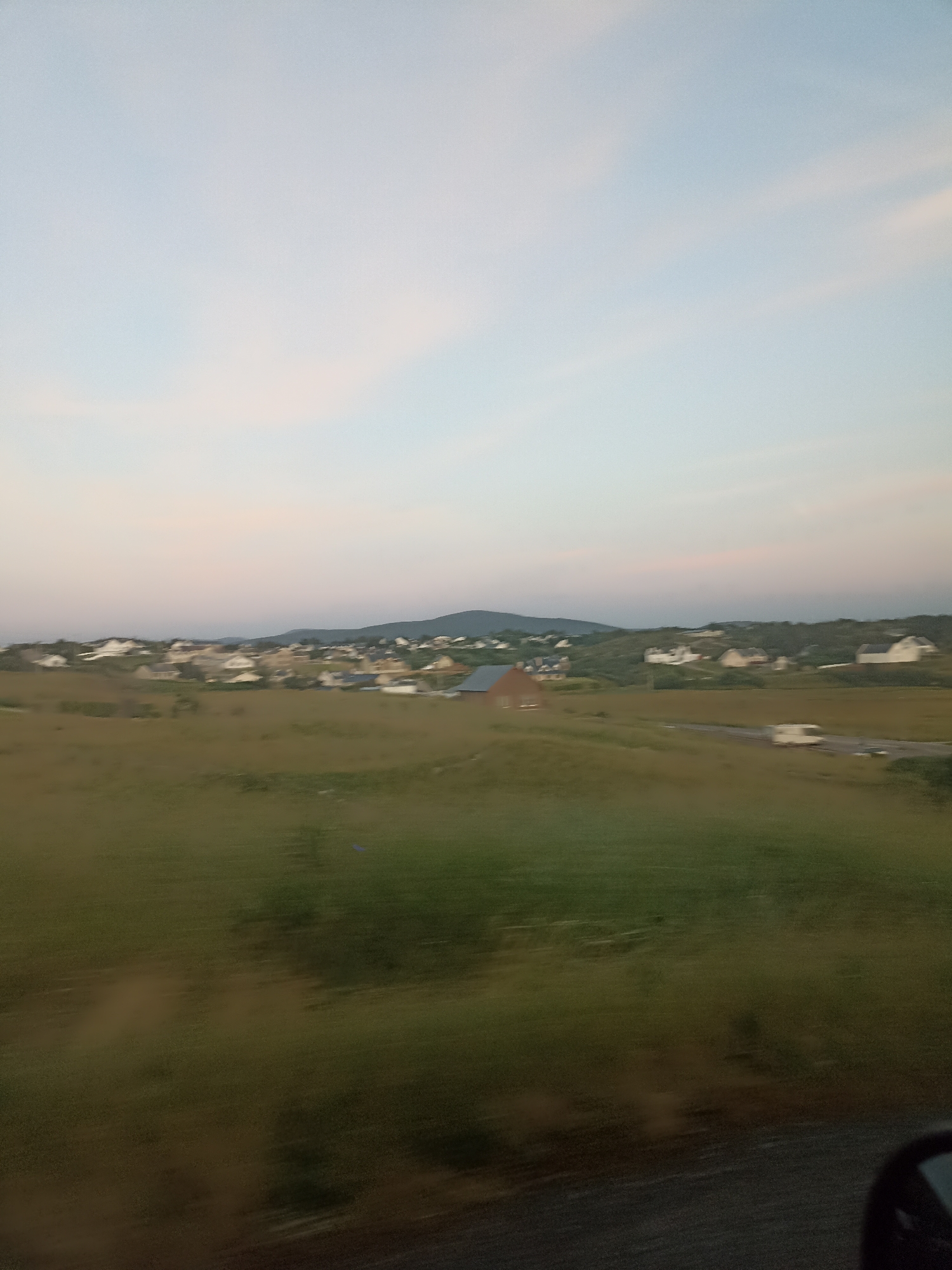 The image captures a rural landscape in Ireland at dusk. The sky is a soft gradient of pale blue and pink, with some wispy clouds. In the mid-ground, a cluster of houses with white and grey roofs are nestled on a gentle slope, appearing to be part of a small village or settlement. Some of the houses are visible from the side, while others are seen from the front, and they are spaced out across the hillside. A road runs horizontally through the scene, with a white campervan or RV moving along it. The foreground is dominated by a grassy field, with the grass appearing slightly blurred, suggesting movement in the camera. Behind the houses, rolling hills covered in vegetation form the background. The overall impression is one of a peaceful, serene evening in the Irish countryside.