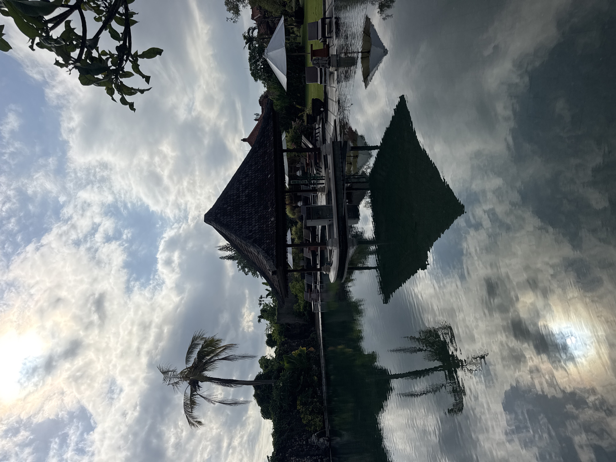 The image shows a water reflection of a tropical landscape, likely taken in the Kuta Selatan area of Indonesia. Dominating the composition is a reflection in the water of a thatched-roof structure, likely a gazebo, with seating underneath. Nearby, a white parasol is also reflected. Lush tropical vegetation surrounds the structure. A palm tree stands tall against a backdrop of partly cloudy sky. The sun, partially obscured, casts a bright glow through the clouds, also reflected in the water. The image has been rotated 180 degrees.