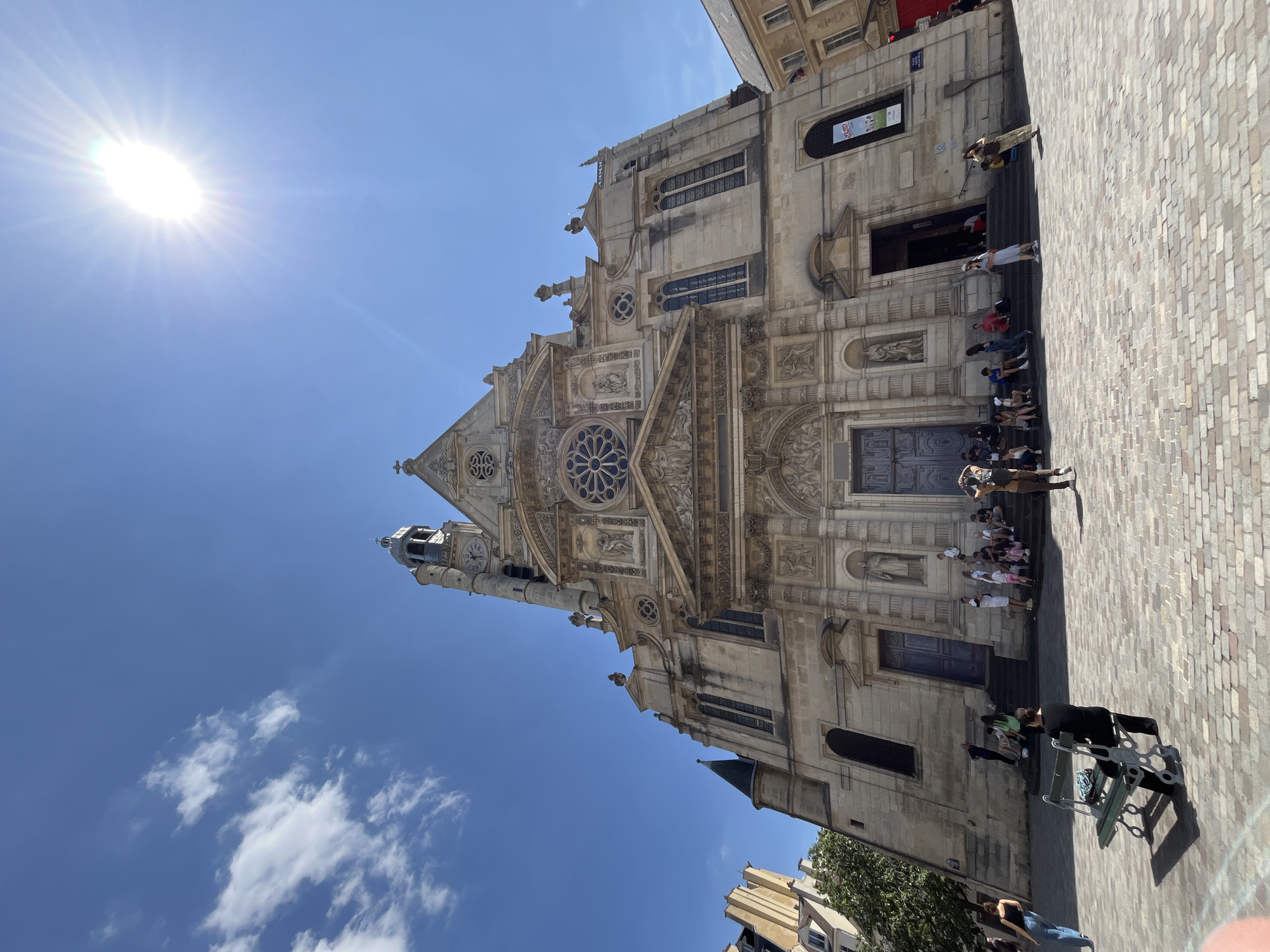 The image shows a low-angle, wide shot of the Church of Saint-Eustache in Paris, France, on a sunny day. The sky is a vibrant blue with a few scattered white clouds and a bright sunbeam emanating from the upper right corner. The church, constructed of light-colored stone, dominates the frame with its ornate facade, featuring a large rose window, intricate sculptures, and a prominent bell tower with a clock. A wide set of stairs leads up to the main entrance, where several people are seated or standing. The cobblestone plaza in front of the church is partially occupied by individuals, some walking, others sitting on benches. In the foreground, a green park bench is positioned to the left, with a person seated on it facing away from the camera. The overall scene depicts a lively public space with a blend of architectural grandeur and everyday human activity.