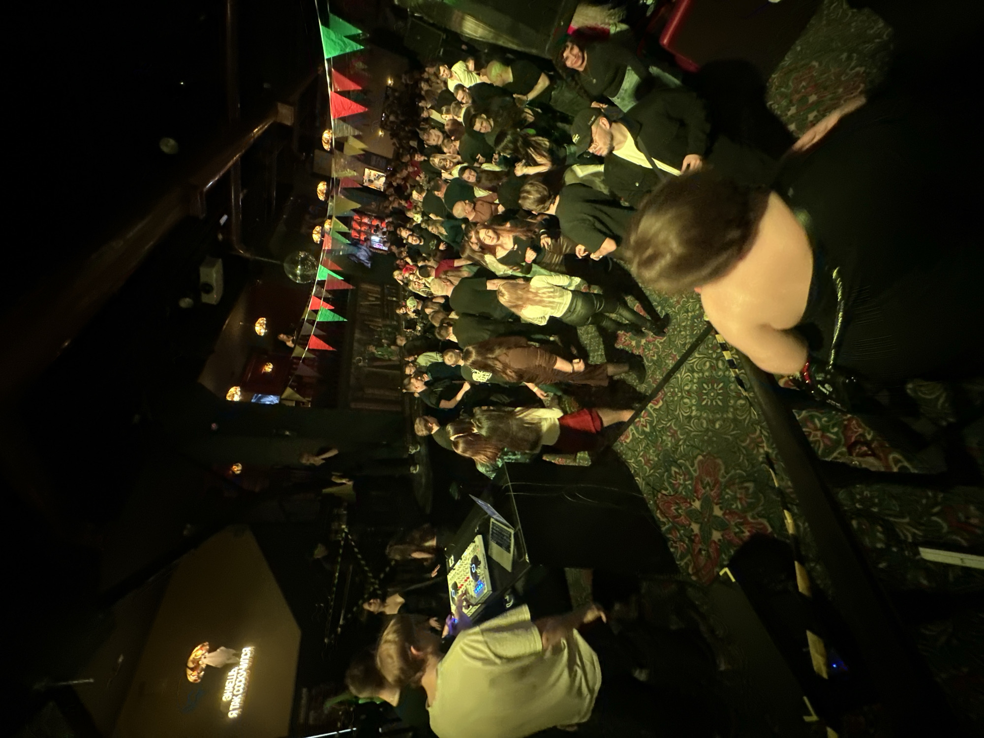 A wide-angle, low-light photograph shows a crowded interior venue, possibly a bar or club. Pennants in green, yellow, and red are strung across the ceiling. People are standing closely packed together, with many facing the camera. A person is standing at a DJ console in the foreground wearing a white shirt. The venue's logo, "ТАК СООБЩИТСЯ" is in the lower left, confirming it is likely in Saratov, Russia. The orientation of the photograph is rotated ninety degrees counter-clockwise.
