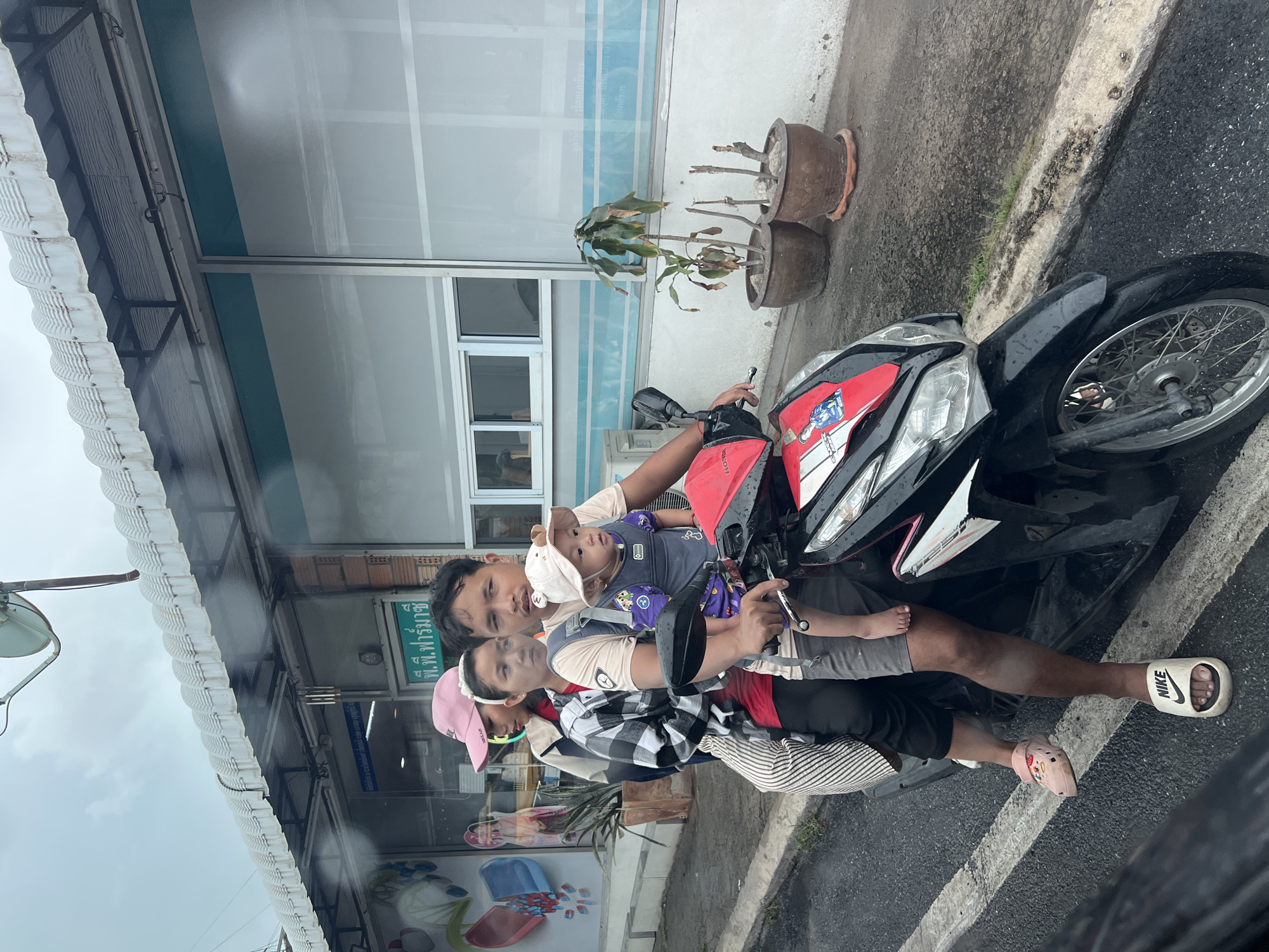 A man, a woman, and a baby are on a black and red motorcycle that is parked along a sidewalk. The man is sitting in the driver's seat with the baby in a carrier in front of him. The woman is sitting behind the man. The man is wearing a white and black Nike sandal. The woman is wearing pink Crocs. The baby is barefoot. The man is holding the handlebars of the motorcycle. There are two planters of dead or dying plants on the sidewalk behind the motorcycle. A building with blue trim is behind the plants. A covered walkway is on the left side of the image. The location is a city in Thailand based on the sign on the building.