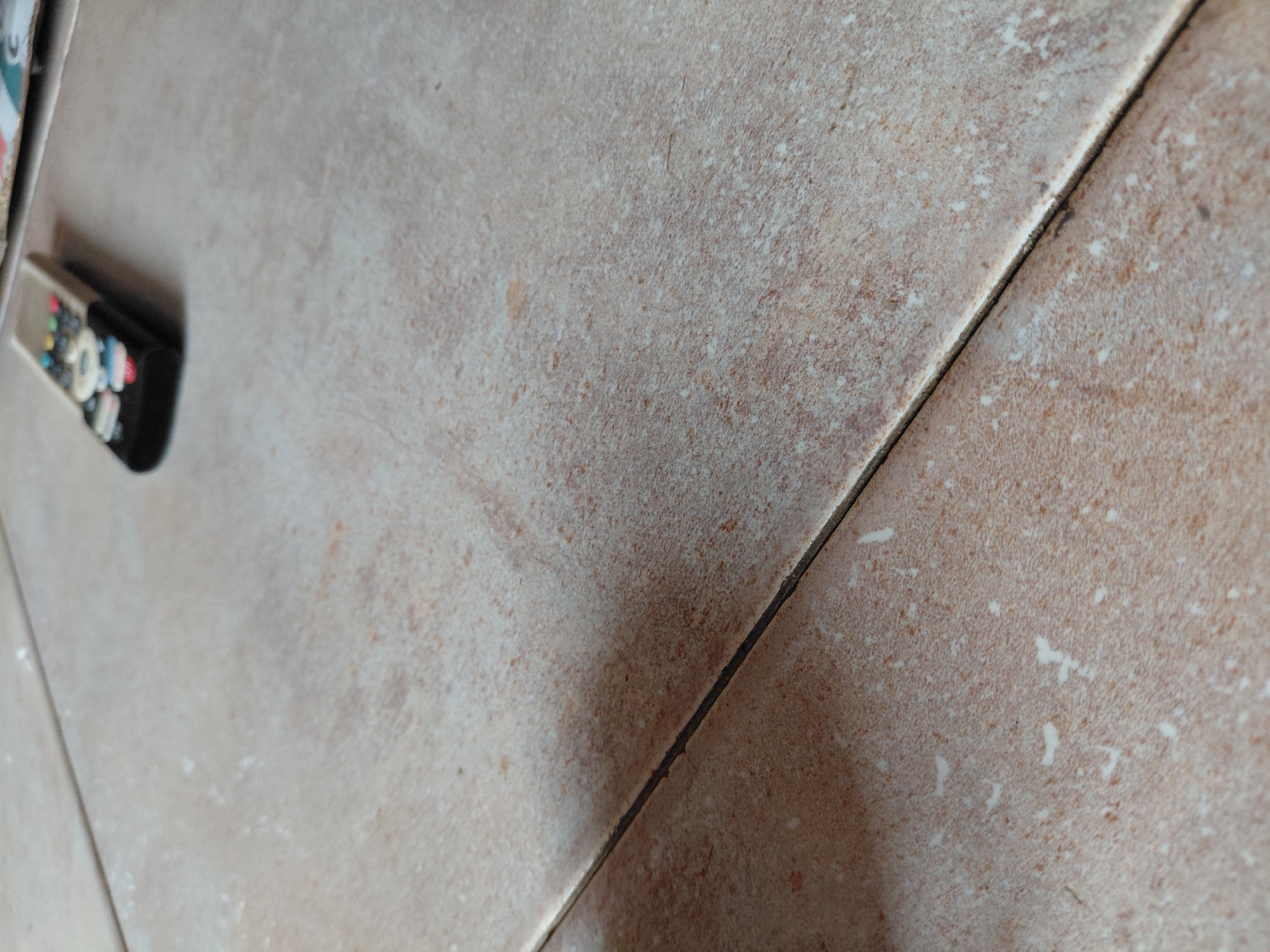 The image displays a close-up, angled perspective of a tiled surface, likely a floor or wall, situated in an indoor setting in Santa Ana, Venezuela. The surface is composed of light brown and beige tiles, possibly ceramic or porcelain, characterized by a speckled, somewhat mottled pattern incorporating lighter and darker tones. Scattered across the tile surfaces are irregular white specks, which could be dust, minor stains, or part of the tile's natural design.
A prominent, dark grout line diagonally traverses the image, separating two of these tiles. This grout line appears dark brown or black and shows some minor irregularities in its consistency.
On the far left side of the frame, partially visible and out of focus, a black remote control rests on the tiled surface. Although blurry, its colorful buttons—including hues of red, green, blue, yellow, and white—are discernible. No specific brand or text on the remote control is clear due to the blur.
The scene depicts a static arrangement of objects; no activity or event is taking place. The lighting suggests ambient indoor conditions. There are no people visible, and no discernible text is present anywhere in the frame. The image primarily focuses on the texture and detail of the tiled surface and the presence of a common household item.