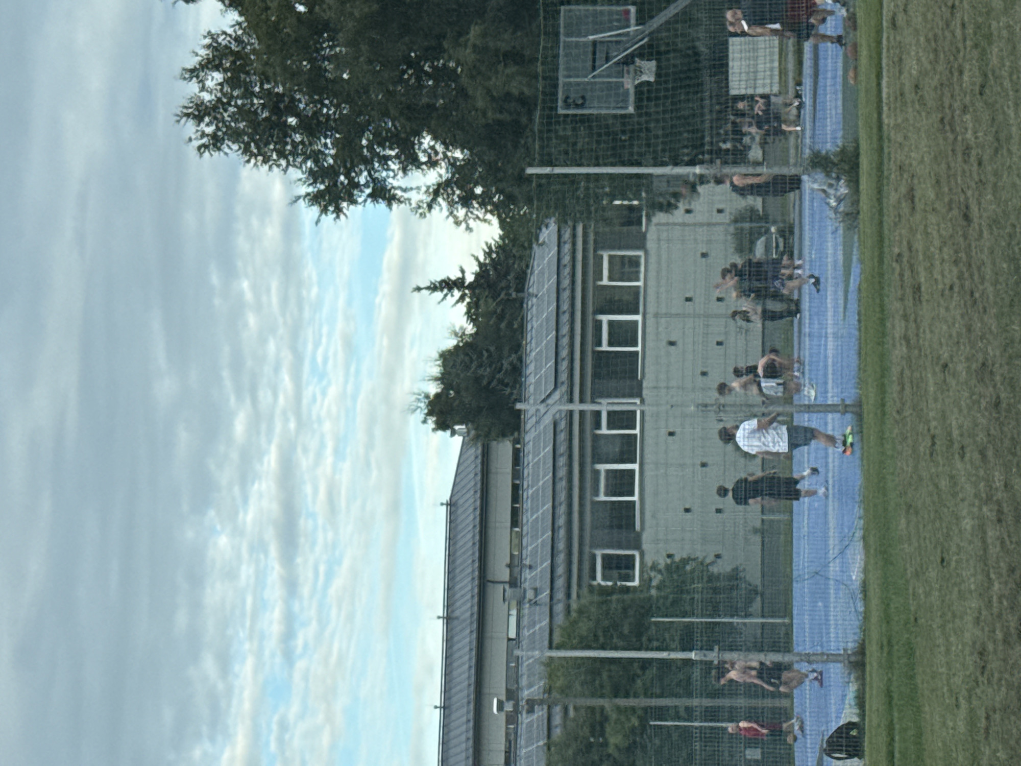 The image shows a basketball court with several individuals playing or observing. The court surface is blue, and a chain-link fence surrounds it. In the background, a modern-looking building with large windows and a dark roof, possibly adorned with solar panels, is visible. Several trees are interspersed between the court and the building, and also to the right of the building. The sky is cloudy with patches of blue visible.
On the court, there are at least ten people. Some are actively engaged in playing basketball, including individuals dribbling, passing, and shooting. Others appear to be standing around, watching the game, or conversing. Their attire consists of casual athletic wear. A basketball hoop with a backboard and net is positioned on the right side of the court.
The foreground is dominated by a grassy area, also enclosed by a fence. The overall scene depicts a casual outdoor activity taking place in an urban or suburban setting. The lighting suggests it is daytime, possibly late afternoon or early evening, given the sky conditions.